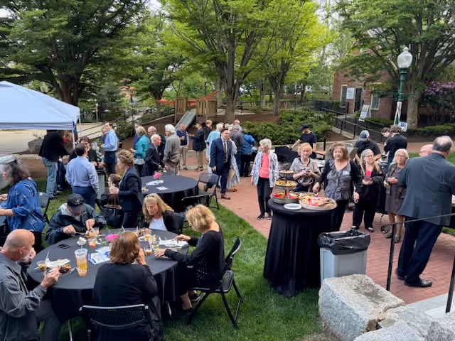 Group of people socializing and eating at an outdoor event with tables, trees, and a brick path.