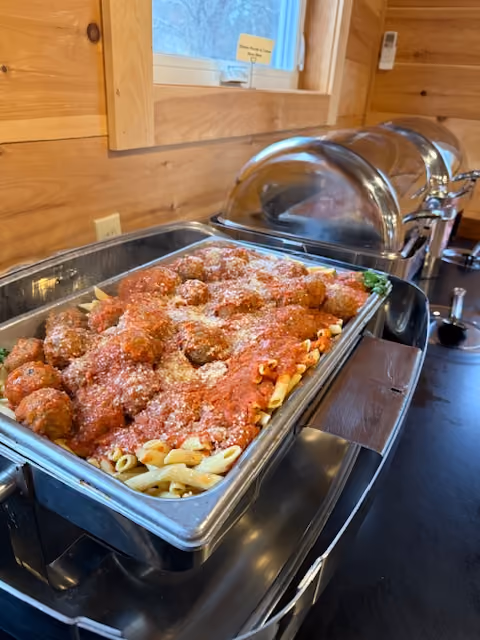 Tray of penne pasta with meatballs and tomato sauce topped with grated cheese in a buffet setup.