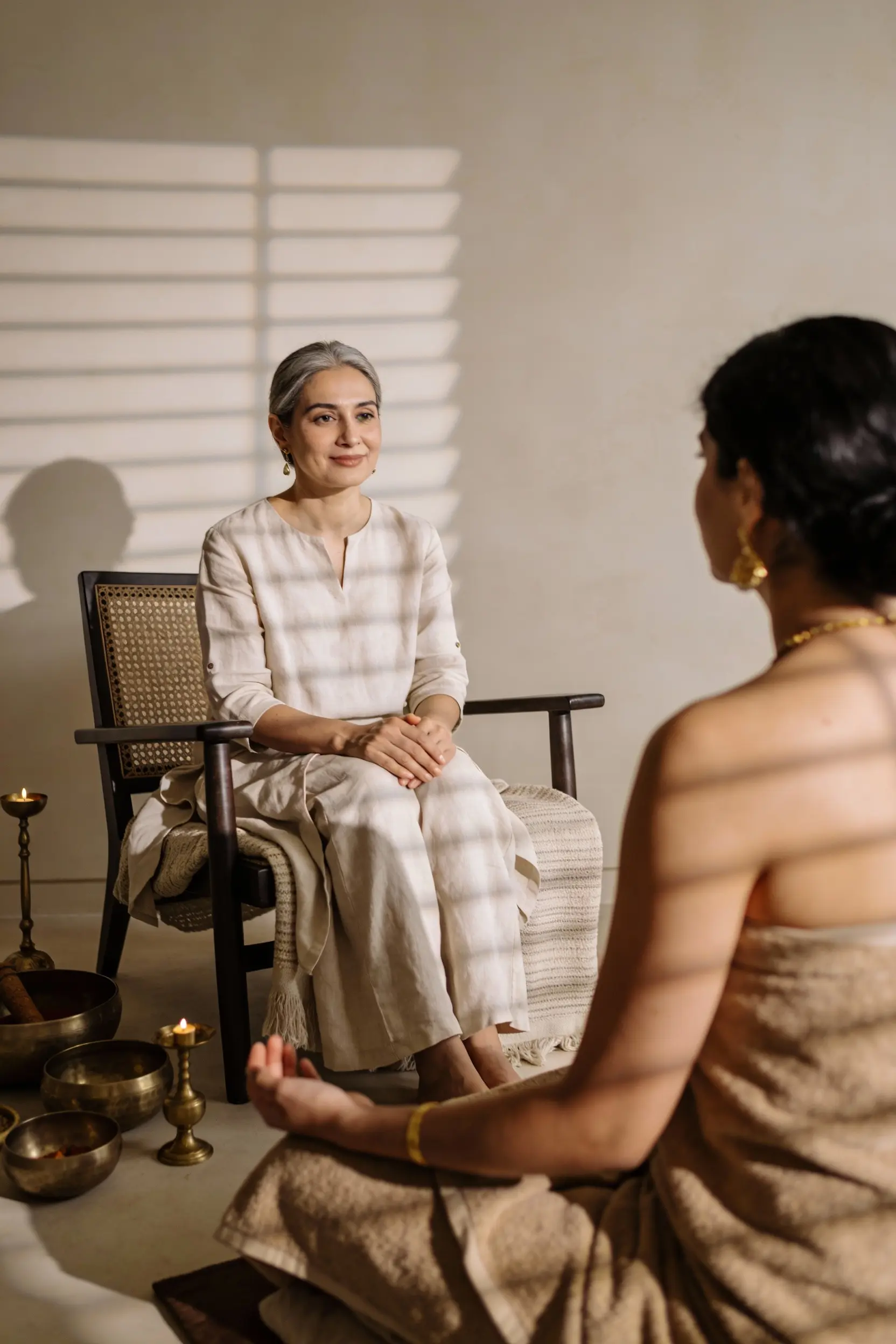 Two women in a serene room, one sitting cross-legged on the floor meditating, the other seated on a chair watching her attentively with soft candlelight around.