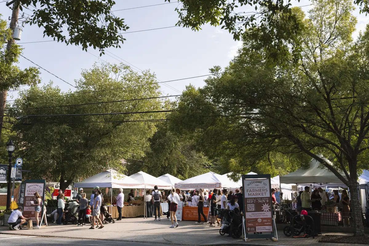Wide view of the Virginia-Highland Farmers Market, with white vendor tents set up under large shade trees and a 'Farmers Market' sign near the entrance.