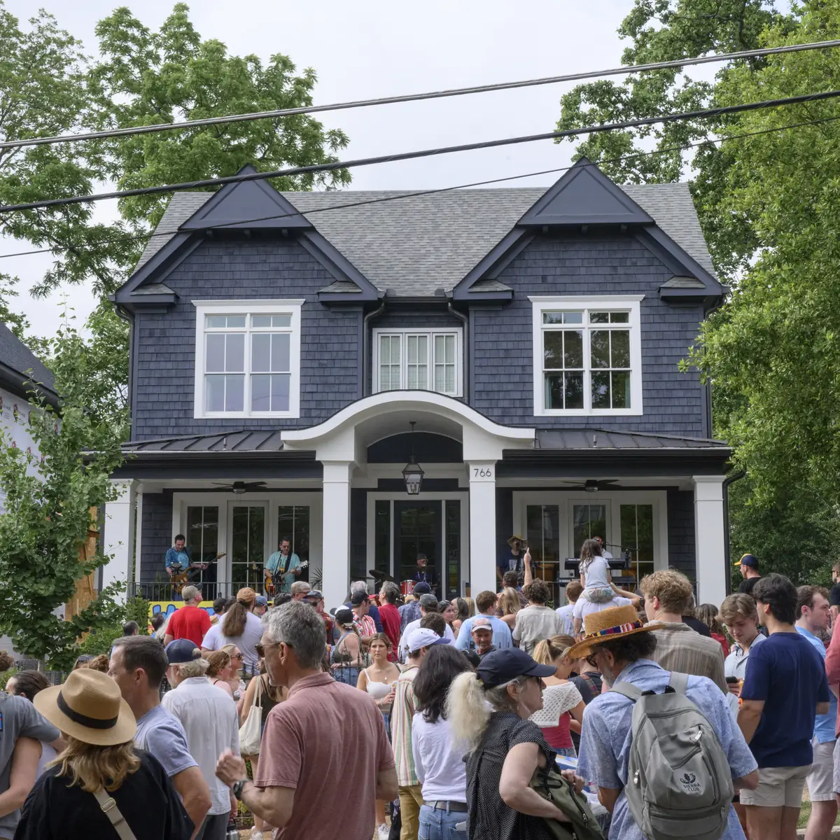 Wide view of a large house with a band playing on the porch and a big crowd gathered on the lawn at Porchfest.