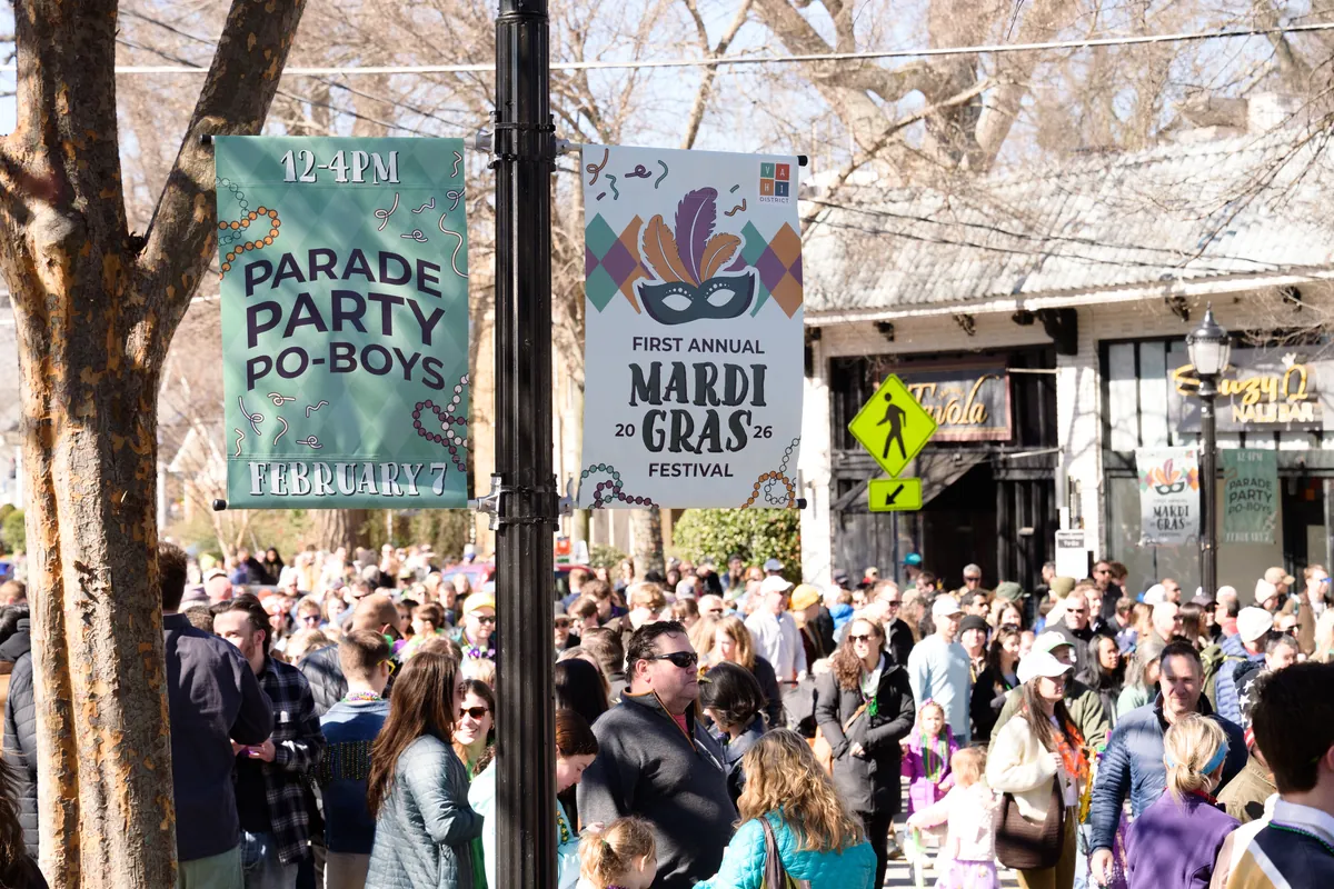 Signs advertising the Parade Party Po-Boys and the First Annual Mardi Gras Festival stand above a large street crowd.