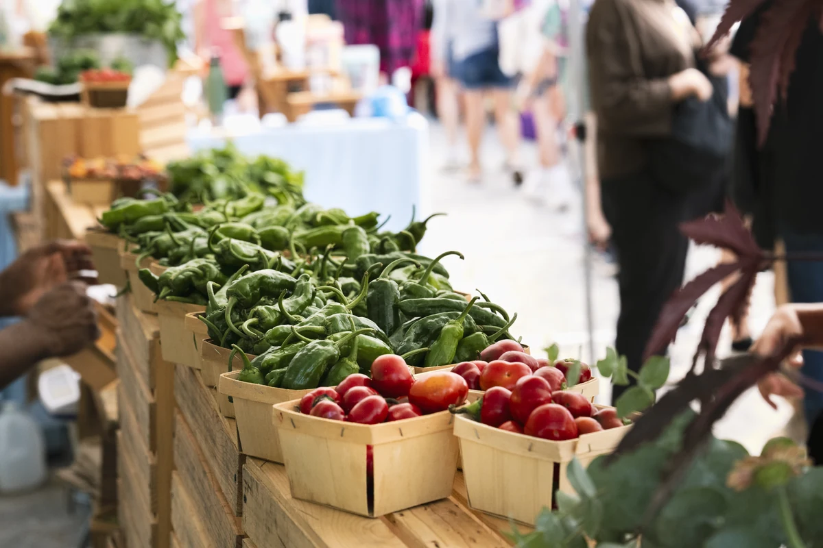 Baskets of green beans and cherry tomatoes displayed on a wooden table at the farmers market, with shoppers browsing in the background.