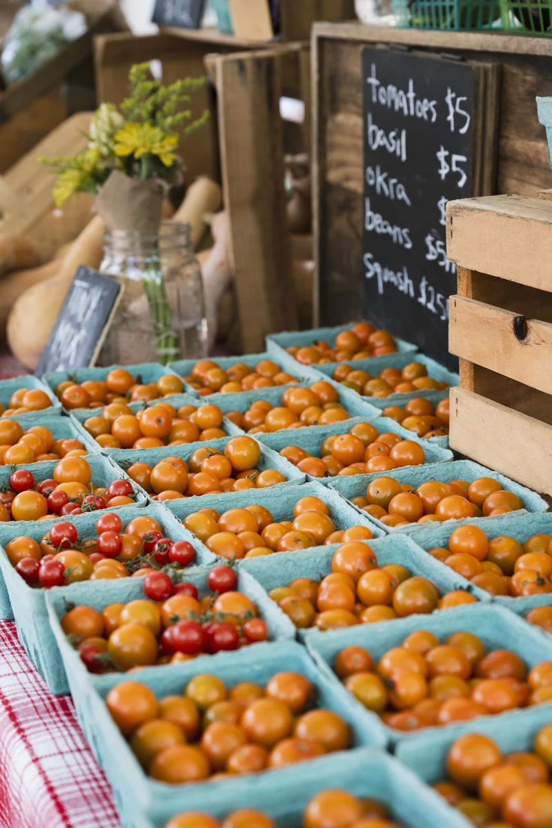 Cartons of orange and red cherry tomatoes for sale at a farmers market stall, with a chalkboard listing prices for tomatoes, basil, okra, beans, and squash.