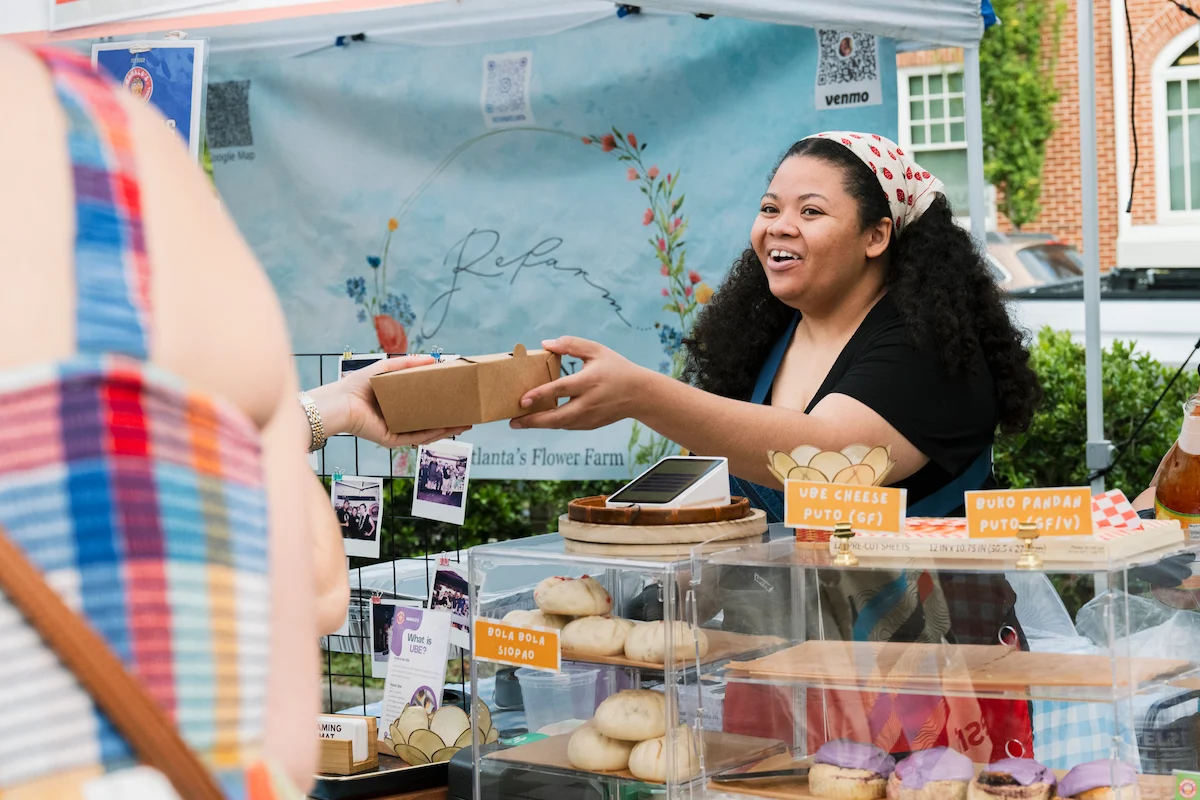 A vendor smiling and handing a box to a customer at a farmers market baked goods stall.