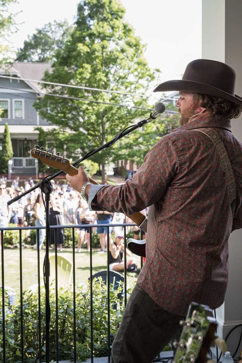 A musician in a cowboy hat plays guitar and sings into a microphone on a porch, with a crowd gathered on the lawn below at Porchfest.