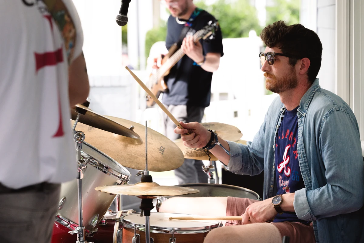A band performs on a porch, with the drummer and guitarist visible mid-song.