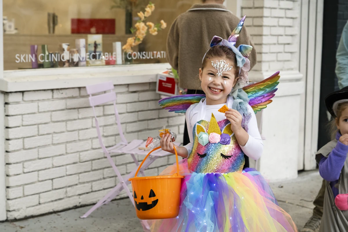 A girl in a unicorn costume with wings holds a jack-o'-lantern bucket outside a shop during Candy Crawl.