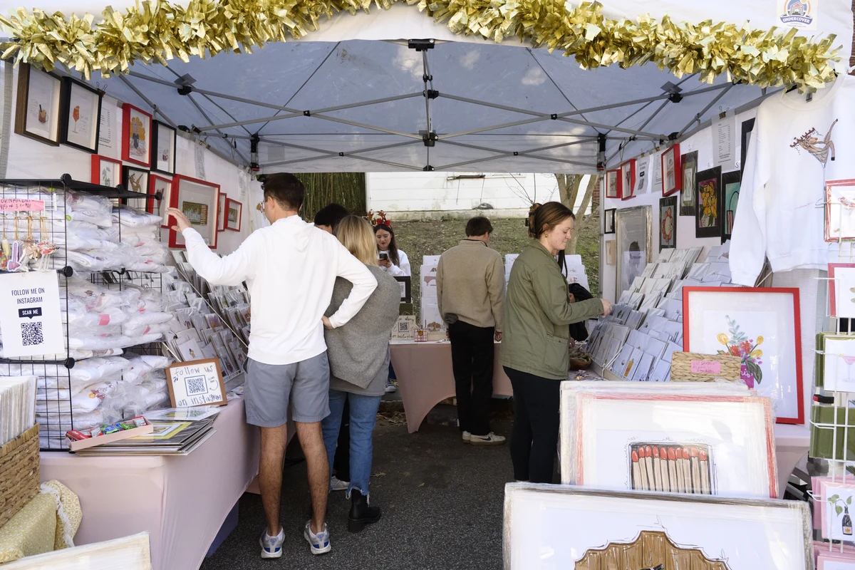 Shoppers browsing art prints and gifts inside a vendor tent decorated with gold garland.