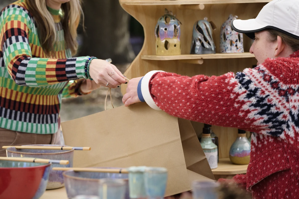 Two people wrapping a paper gift bag at a pottery vendor booth, with handmade bowls displayed nearby.