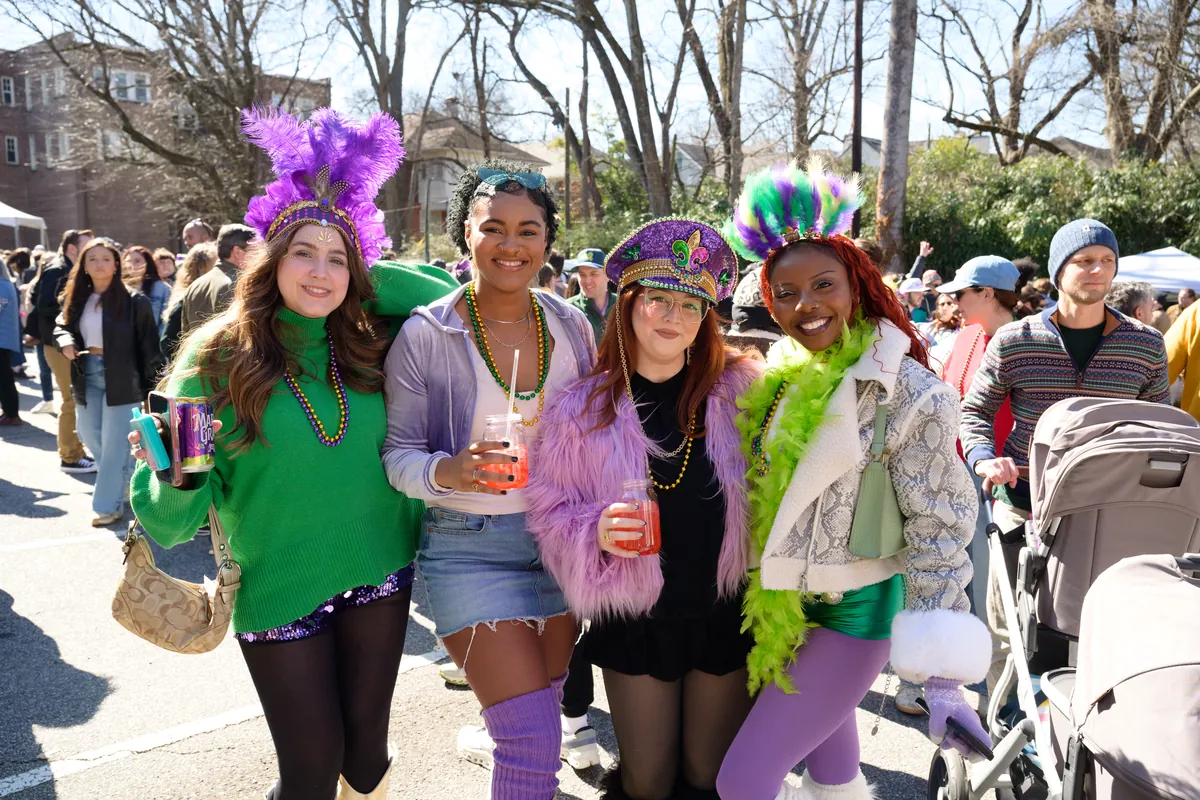 Four women in feathered Mardi Gras headpieces and beads pose together with drinks at the festival.
