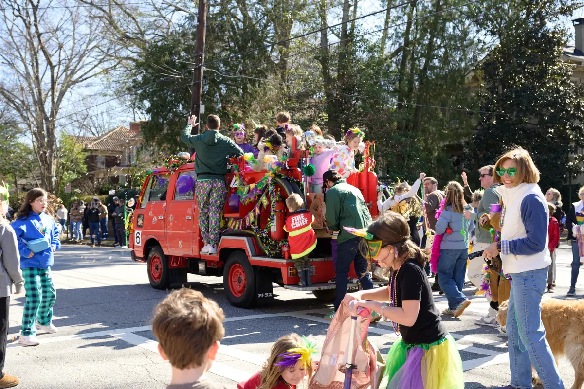 A colorfully decorated parade float made from a fire truck carries riders in Mardi Gras costumes down the street, with a crowd watching.