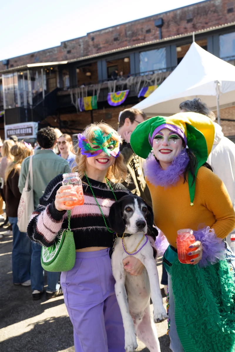 Two women in Mardi Gras masks and costumes pose with a dog wearing beads, holding drinks.