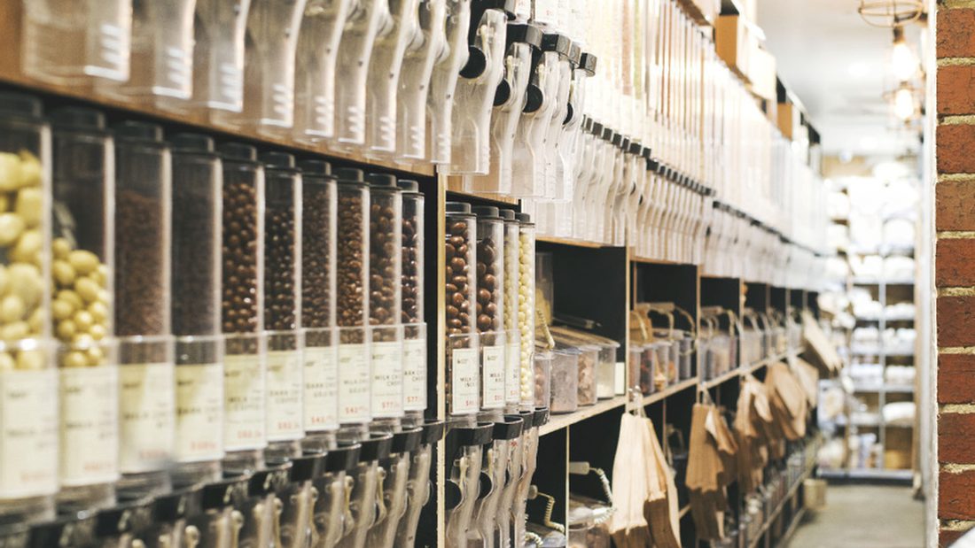 The bulk shelves — two rows of gravity dispensers of nuts, pulses and chocolate, with cloth refill bags hanging beneath and scoops on the shelf.