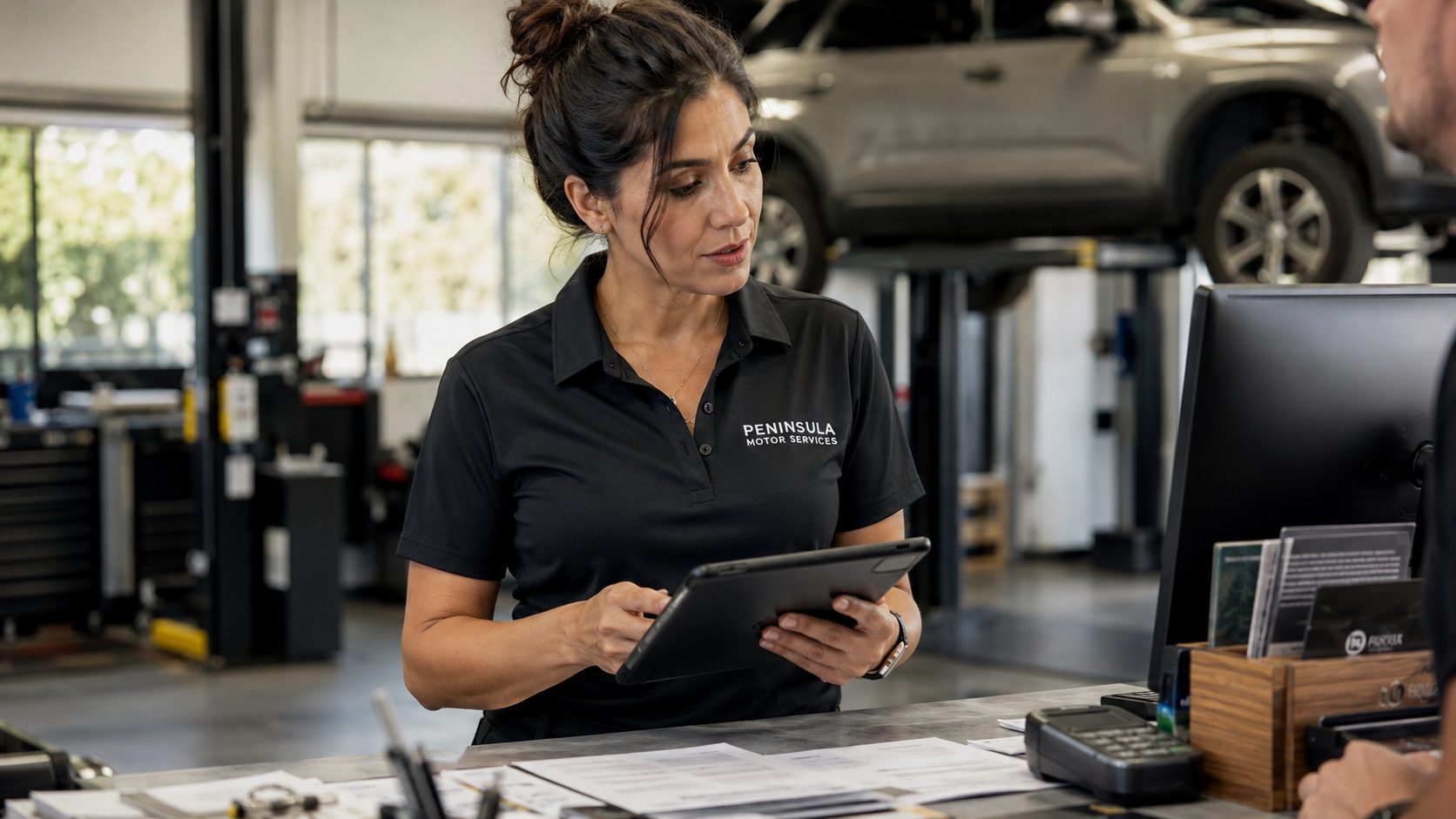Service centre manager reviewing customer data on a tablet at the counter, a vehicle on a lift behind