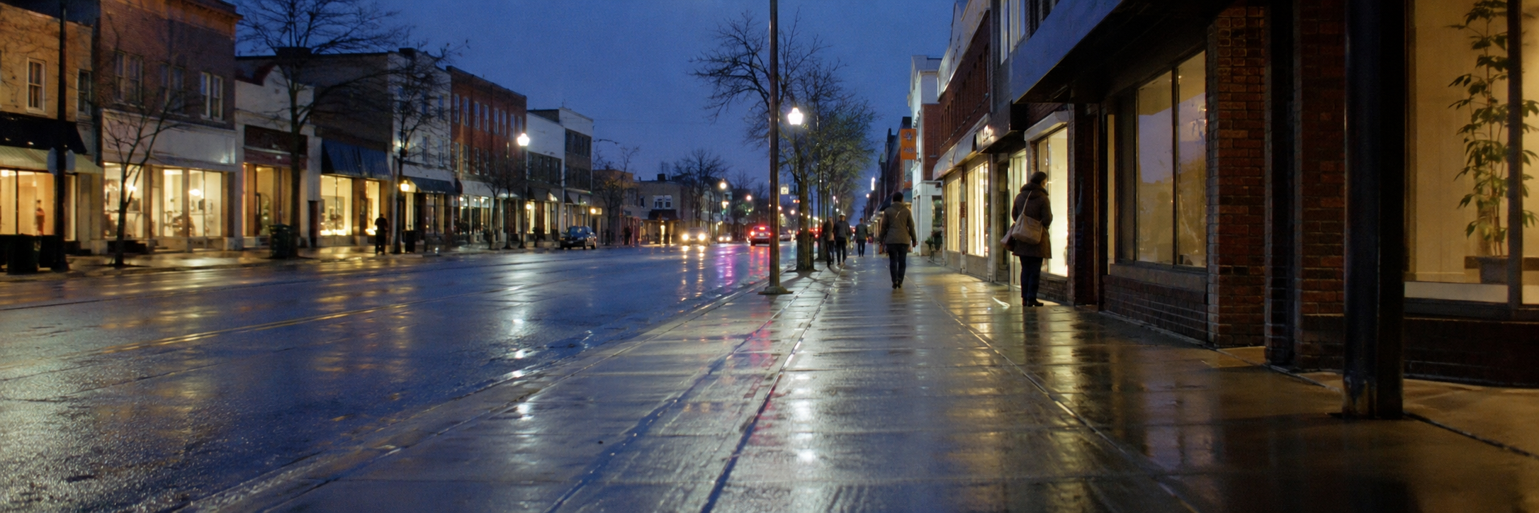 A main street in a small city at dusk, shop windows lit and reflected on wet pavement.