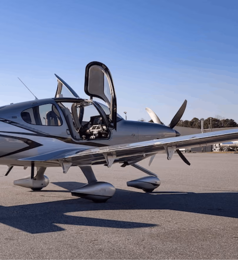 Small silver single-engine airplane with open doors parked on airport tarmac under clear blue sky.