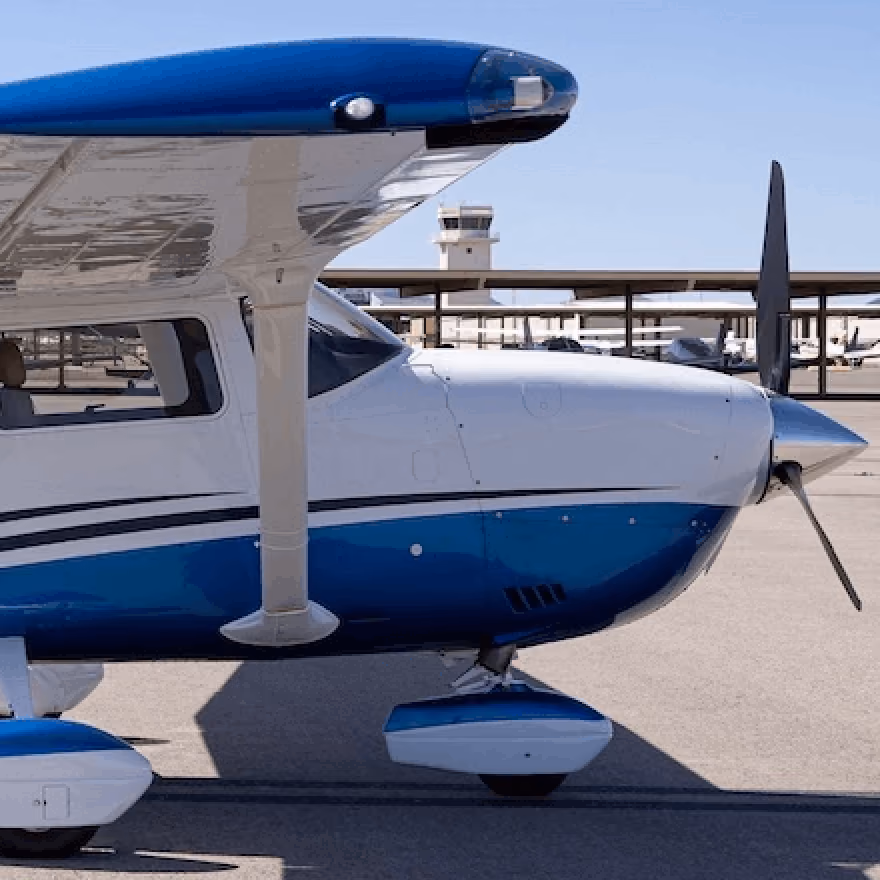 Small white and blue propeller airplane parked on tarmac near airport control tower.