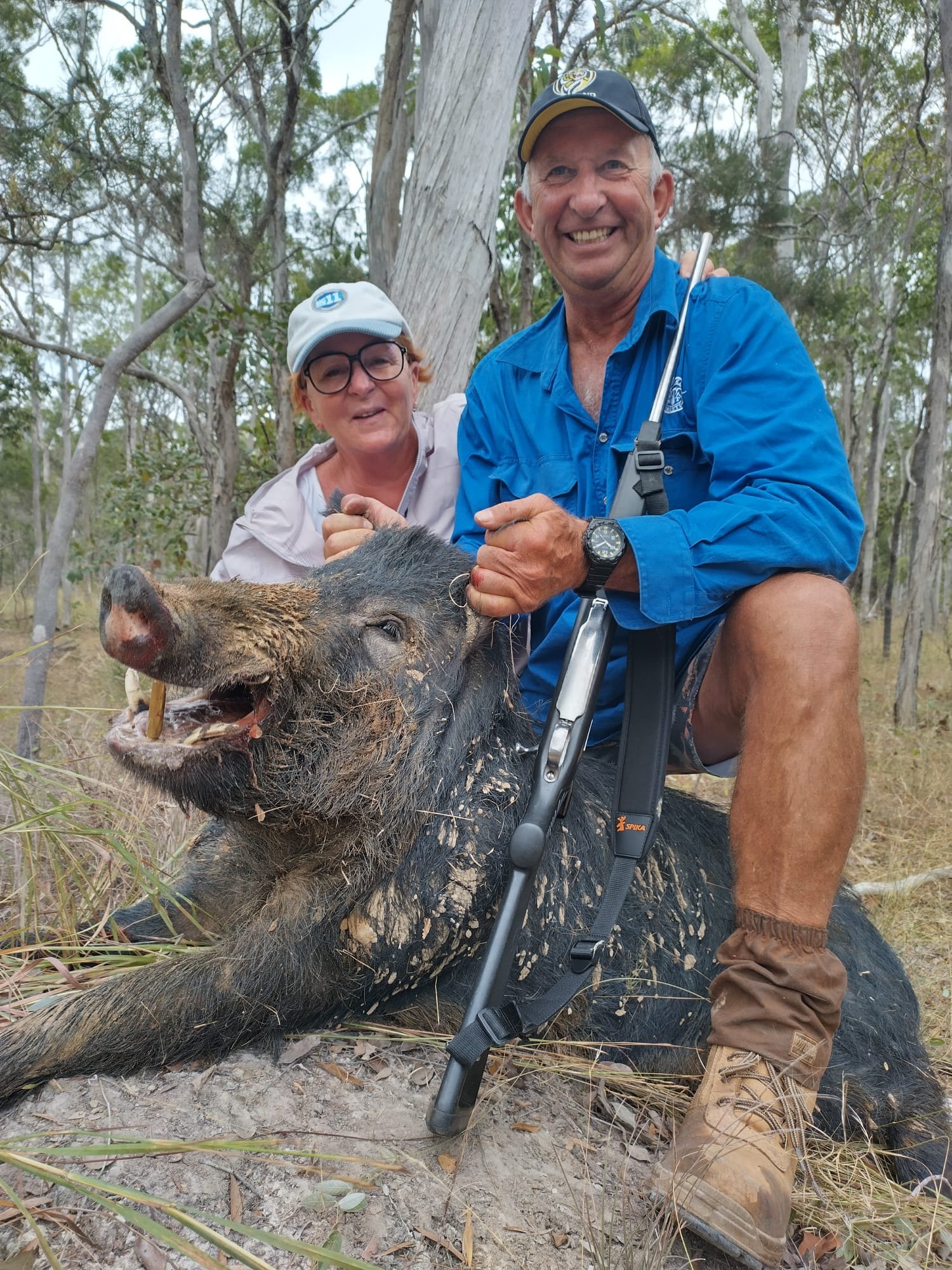 A hunter in a blue shirt kneeling with a rifle beside a large wild boar, with a guest smiling behind