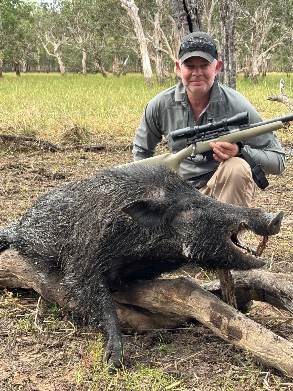 A hunter with a scoped rifle kneeling behind a mud-covered wild boar on a log