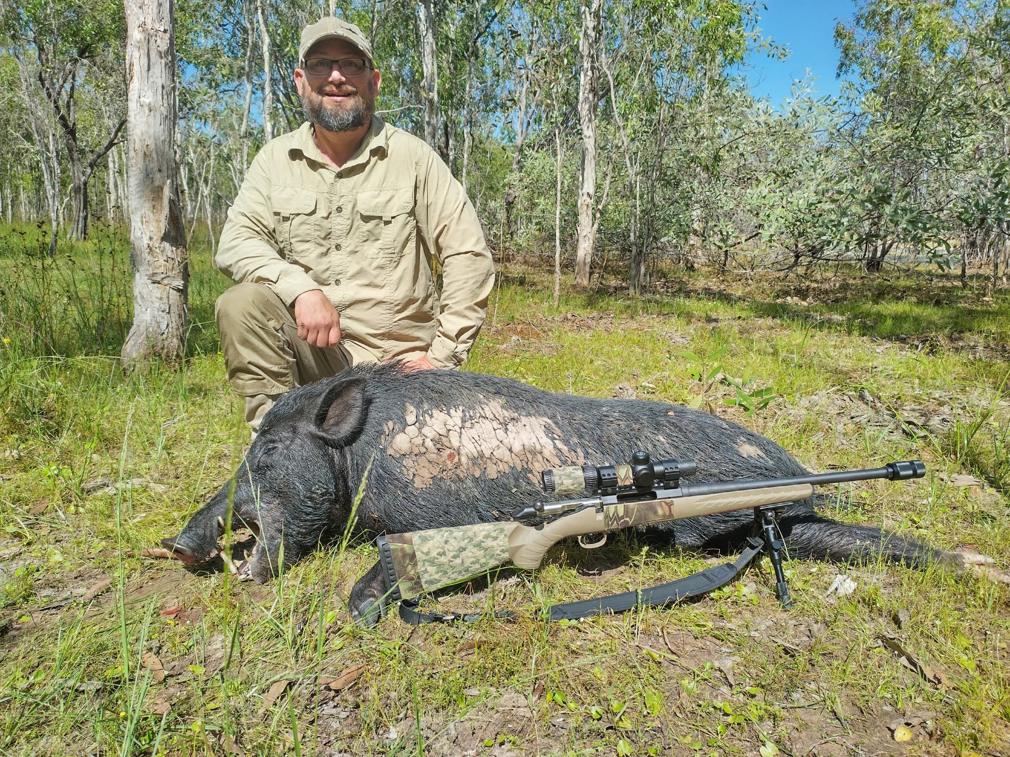 A hunter in khaki kneeling behind a wild boar with a rifle laid alongside