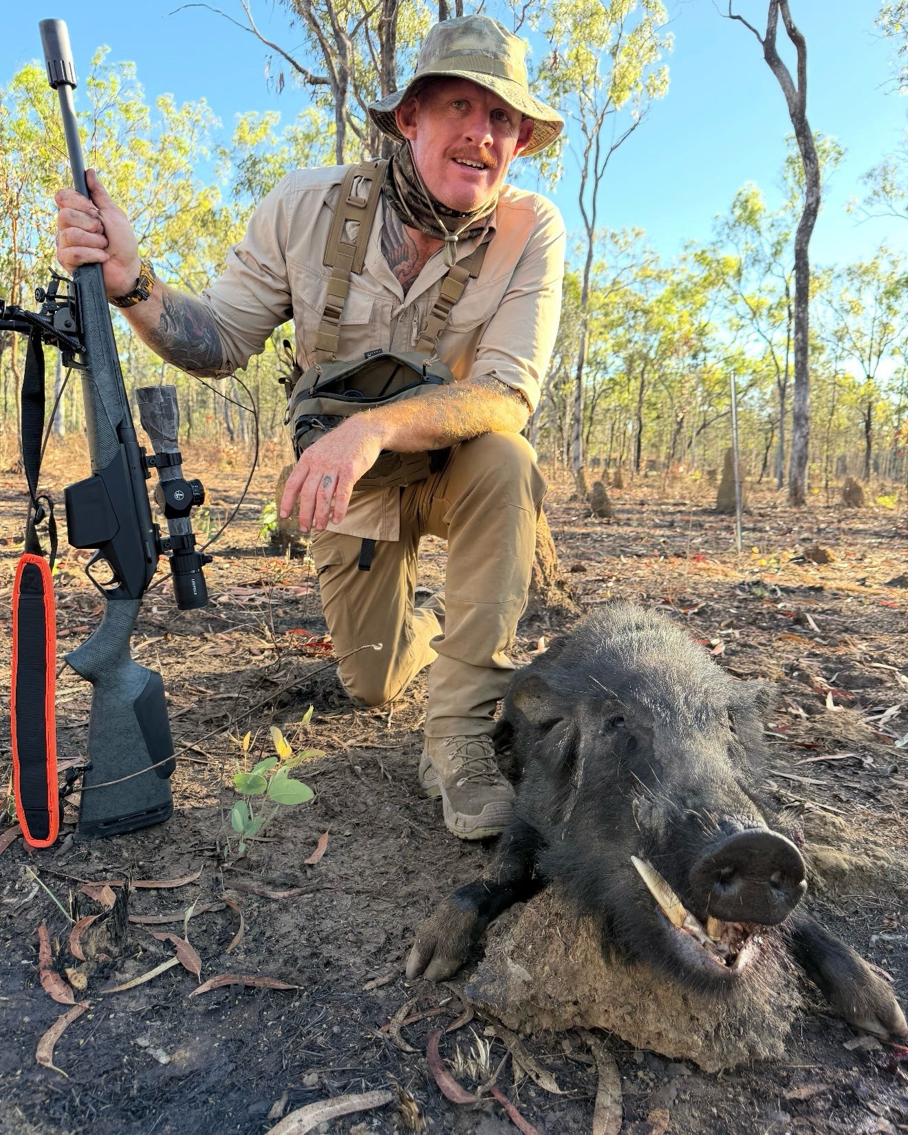 A hunter in field gear kneeling with a rifle behind a boar, with a second boar's head in front