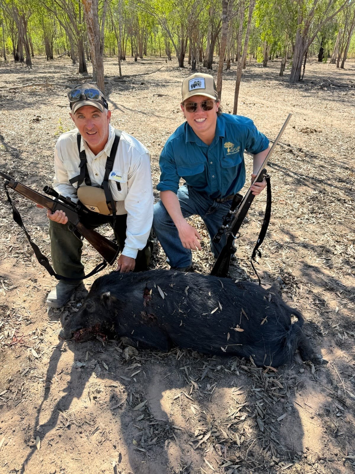 Two hunters with rifles crouching behind a black wild boar on open ground