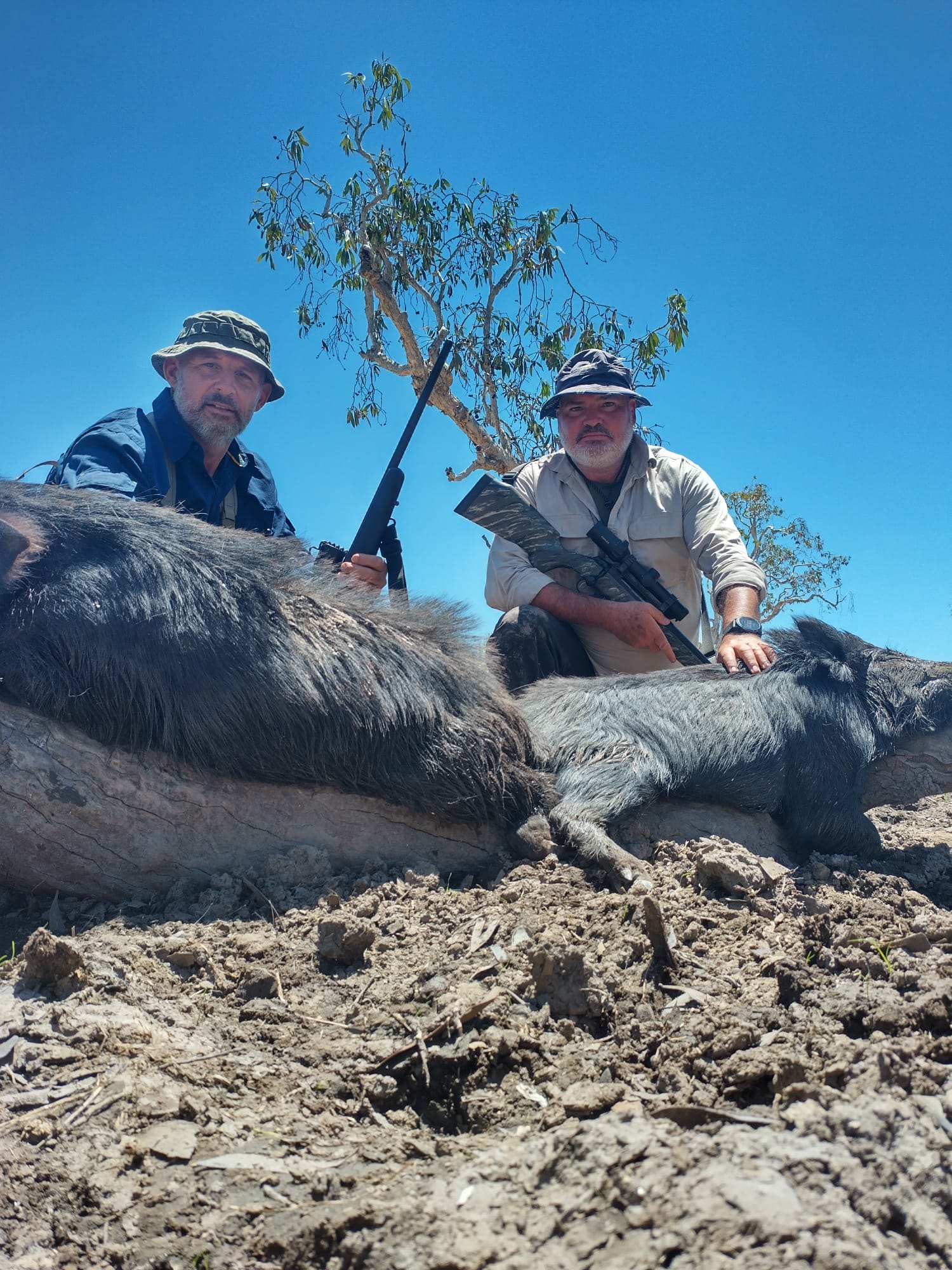 Two hunters in bucket hats crouched behind a pile of wild boar under a blue sky