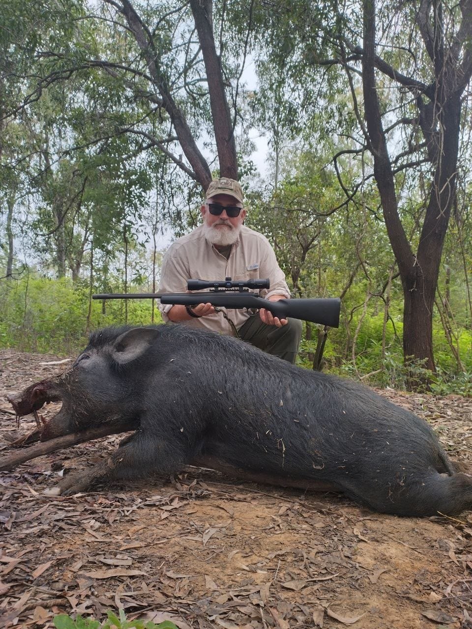 A hunter with a scoped rifle sitting behind a black wild boar in open forest