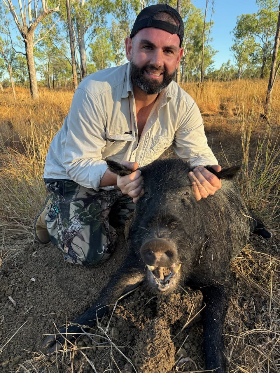 A smiling hunter in a light shirt crouching behind a wild boar