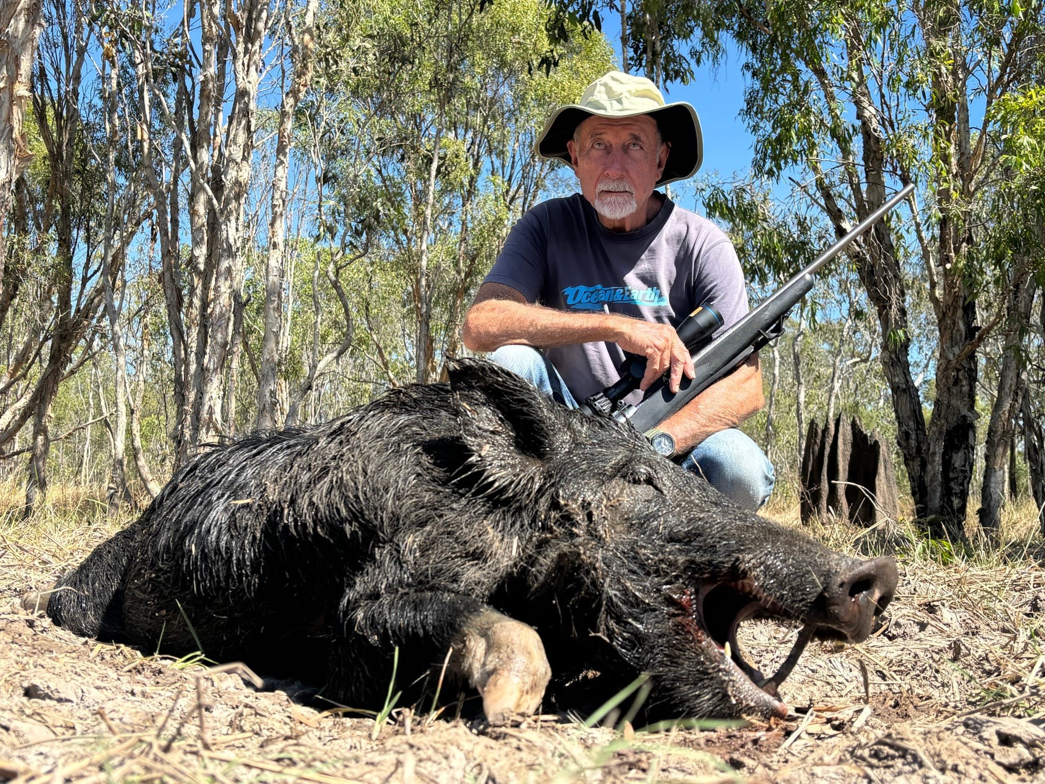 A hunter in a bucket hat crouching with a rifle behind a large wild boar