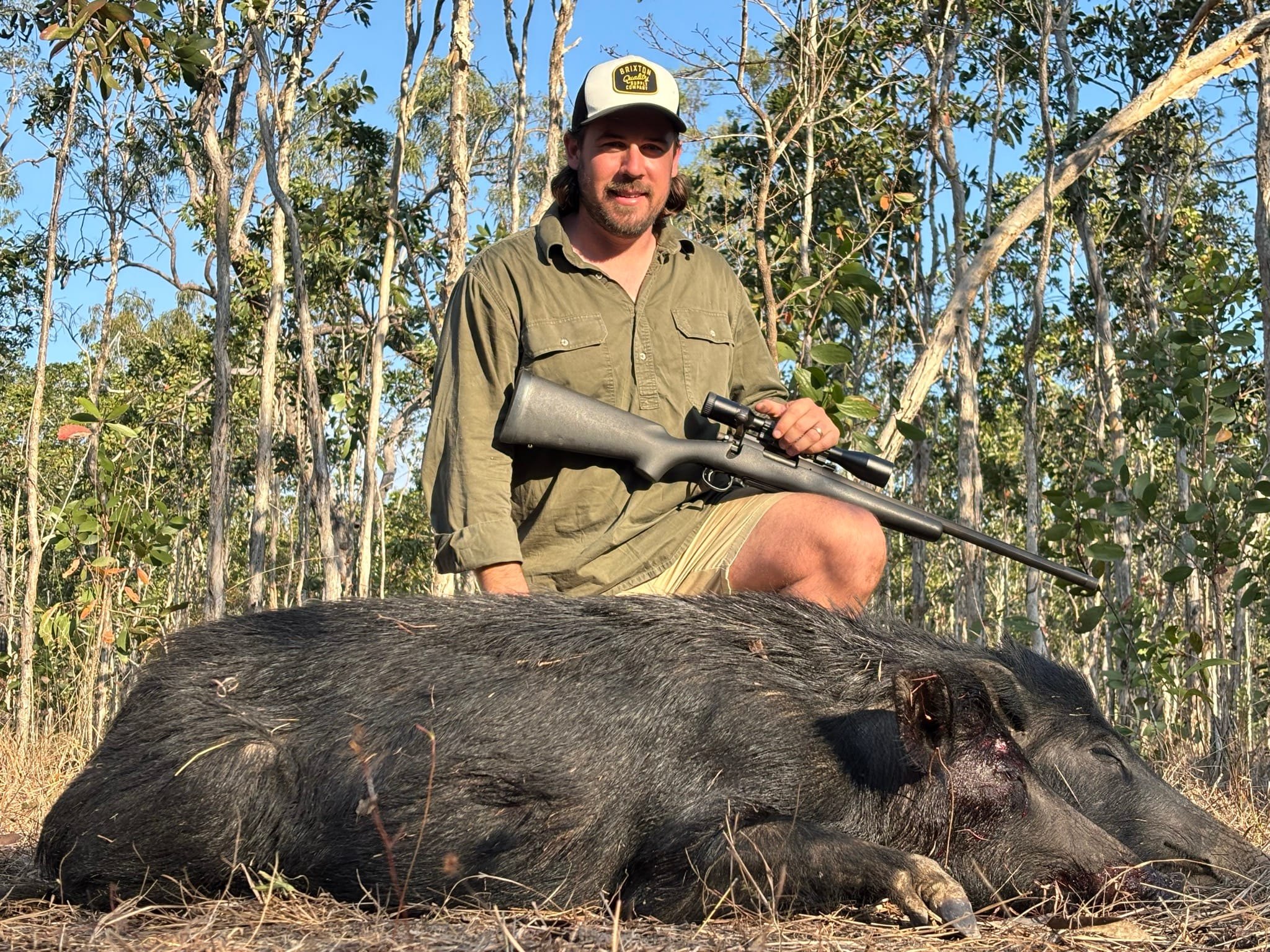A hunter in an olive shirt with a rifle across the knees behind a wild boar