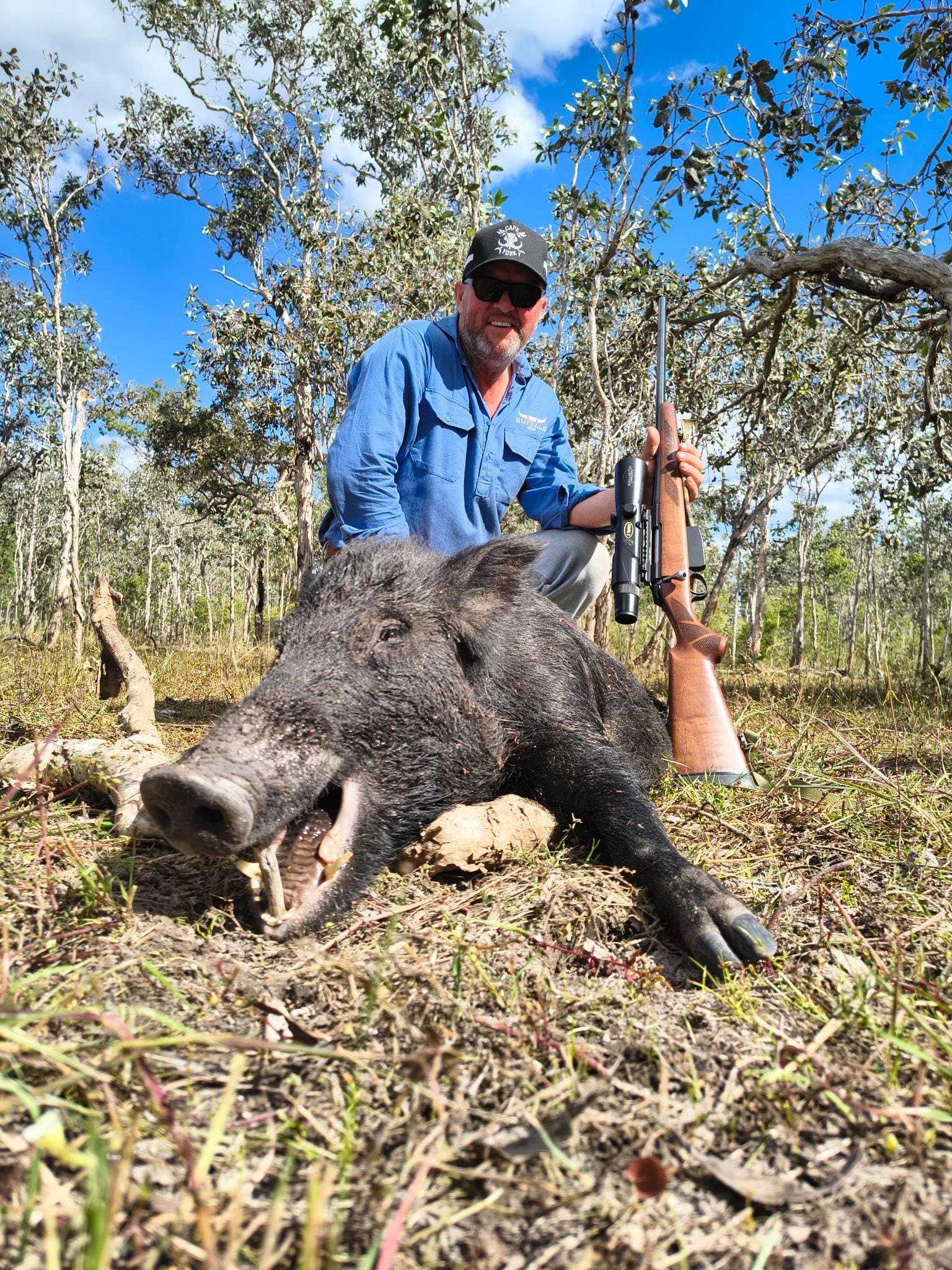 A hunter in a blue shirt and cap with a rifle beside a wild boar in open country