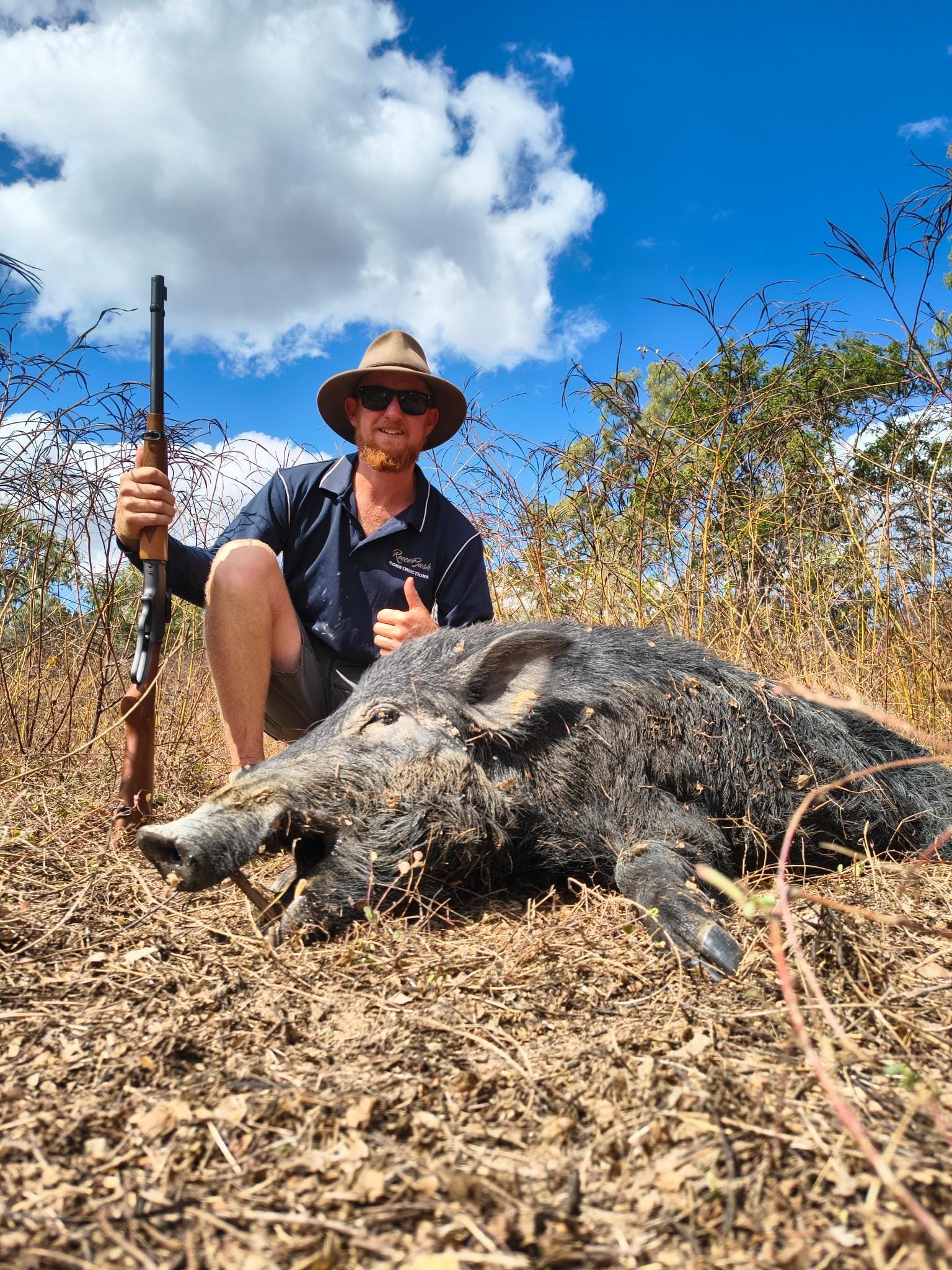 A hunter in a wide-brim hat and sunglasses giving a thumbs up behind a wild boar