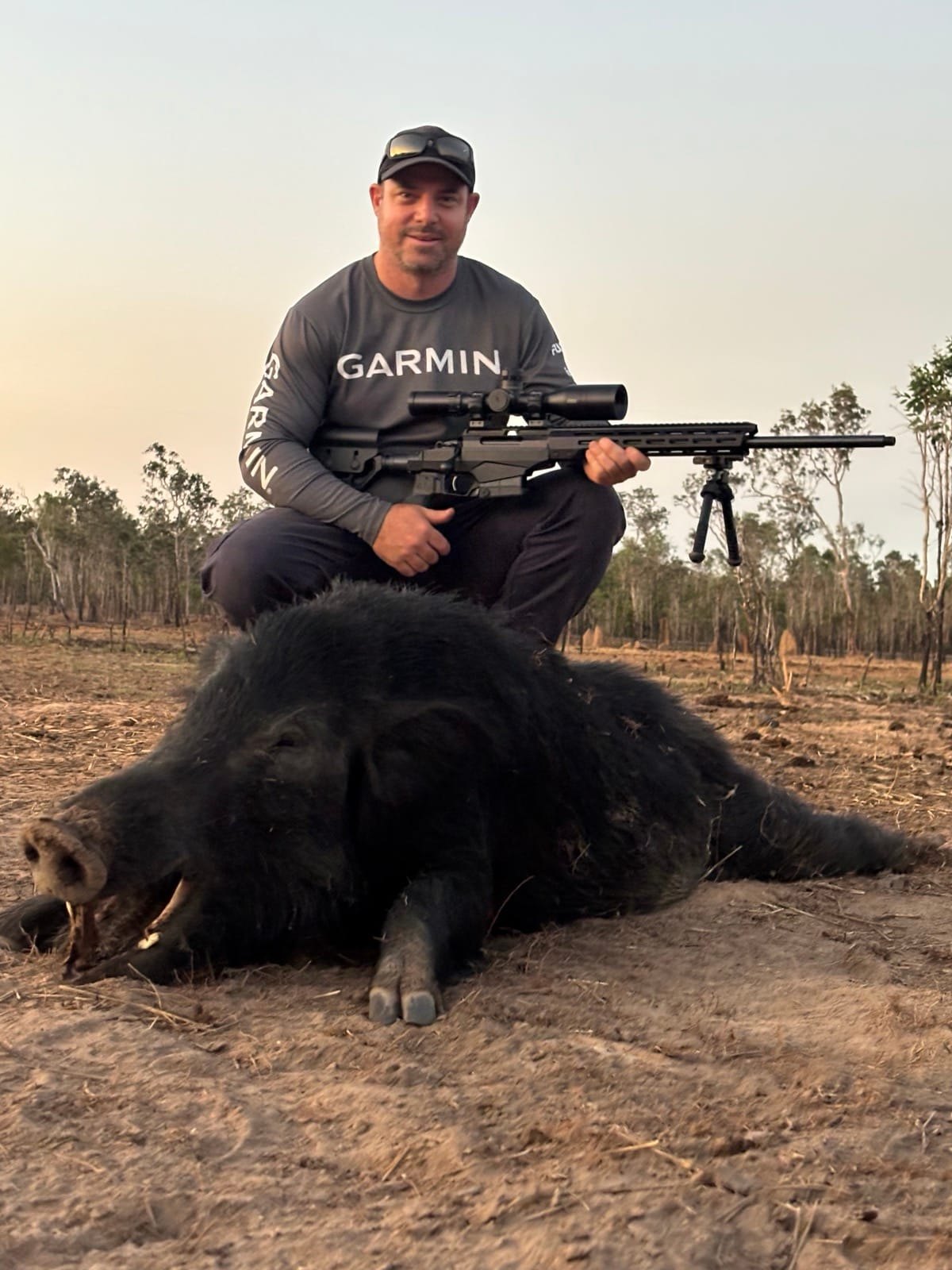 A hunter with a bipod rifle crouching behind a large wild boar at dusk