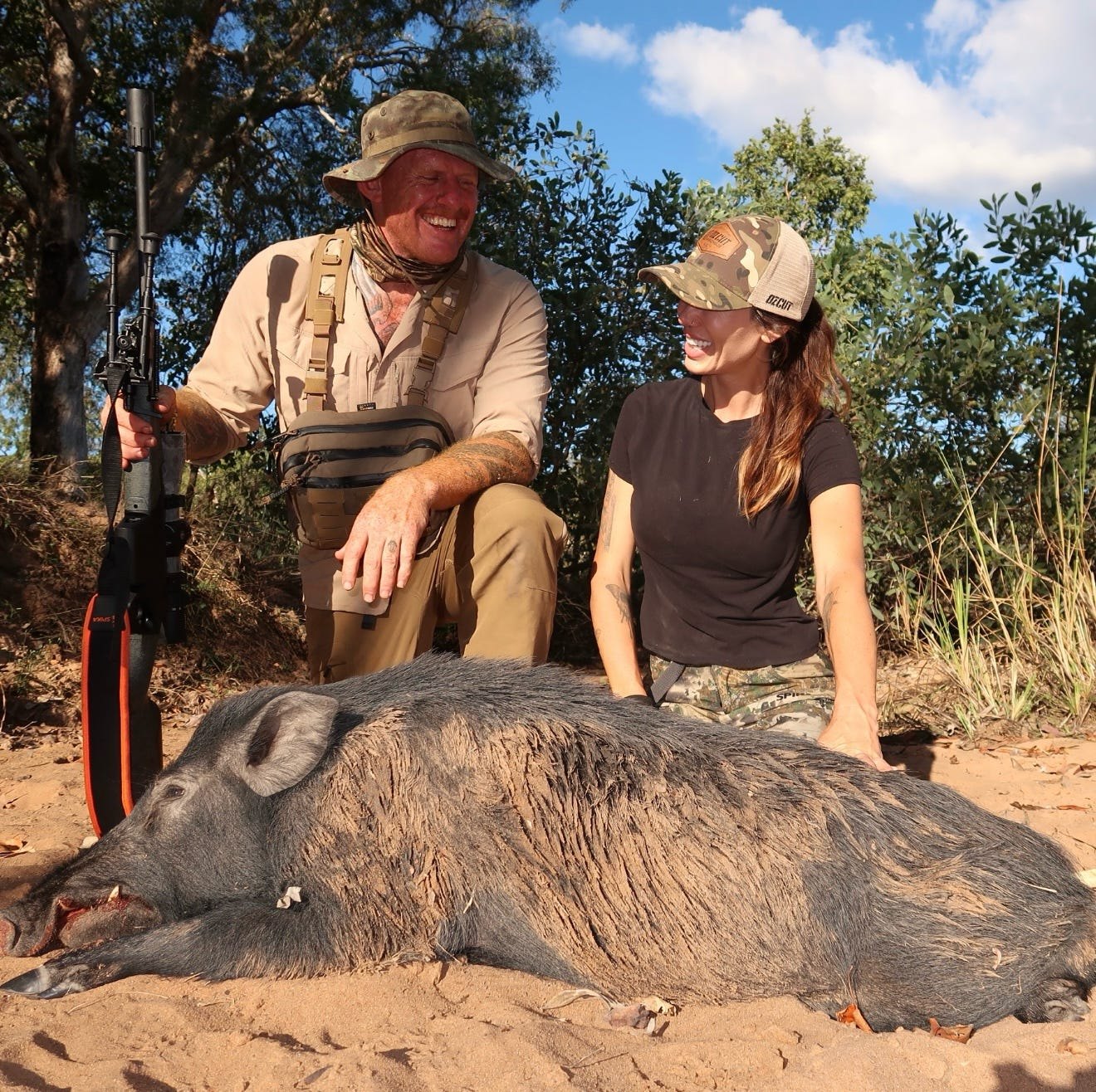 A hunter and a guest crouching behind a boar lying on sandy ground