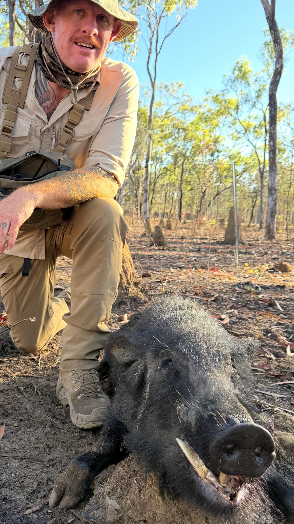 A hunter in a wide-brim hat crouching beside a wild boar, photographed from ground level