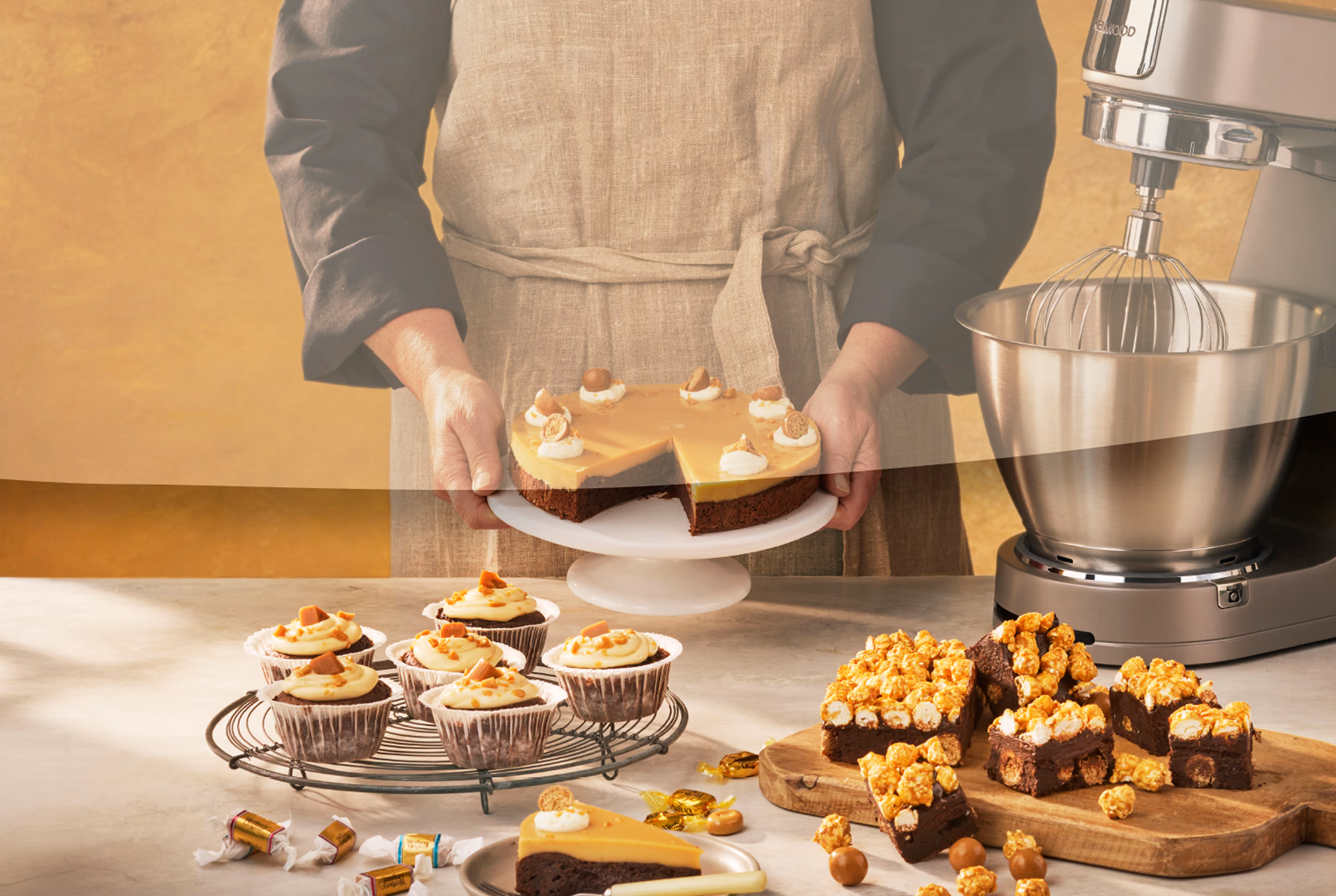 Person holding a caramel-topped cake with cupcakes and caramel popcorn brownies on the table.