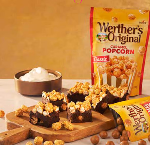 Squares of chocolate brownies topped with Werther's caramel popcorn on wooden board with popcorn bags nearby.