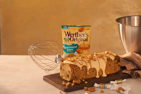 Loaf cake with caramel glaze and nuts on cutting board, Werther's Original bag and whisk nearby.