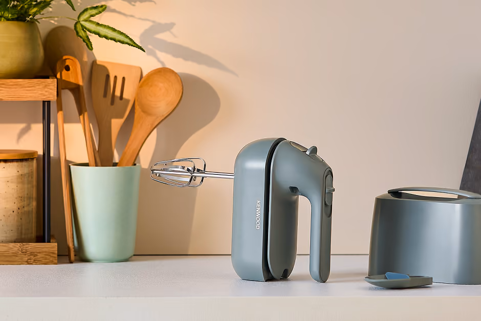 Gray Kenwood hand mixer with beaters on a kitchen counter next to wooden utensils and a plant.