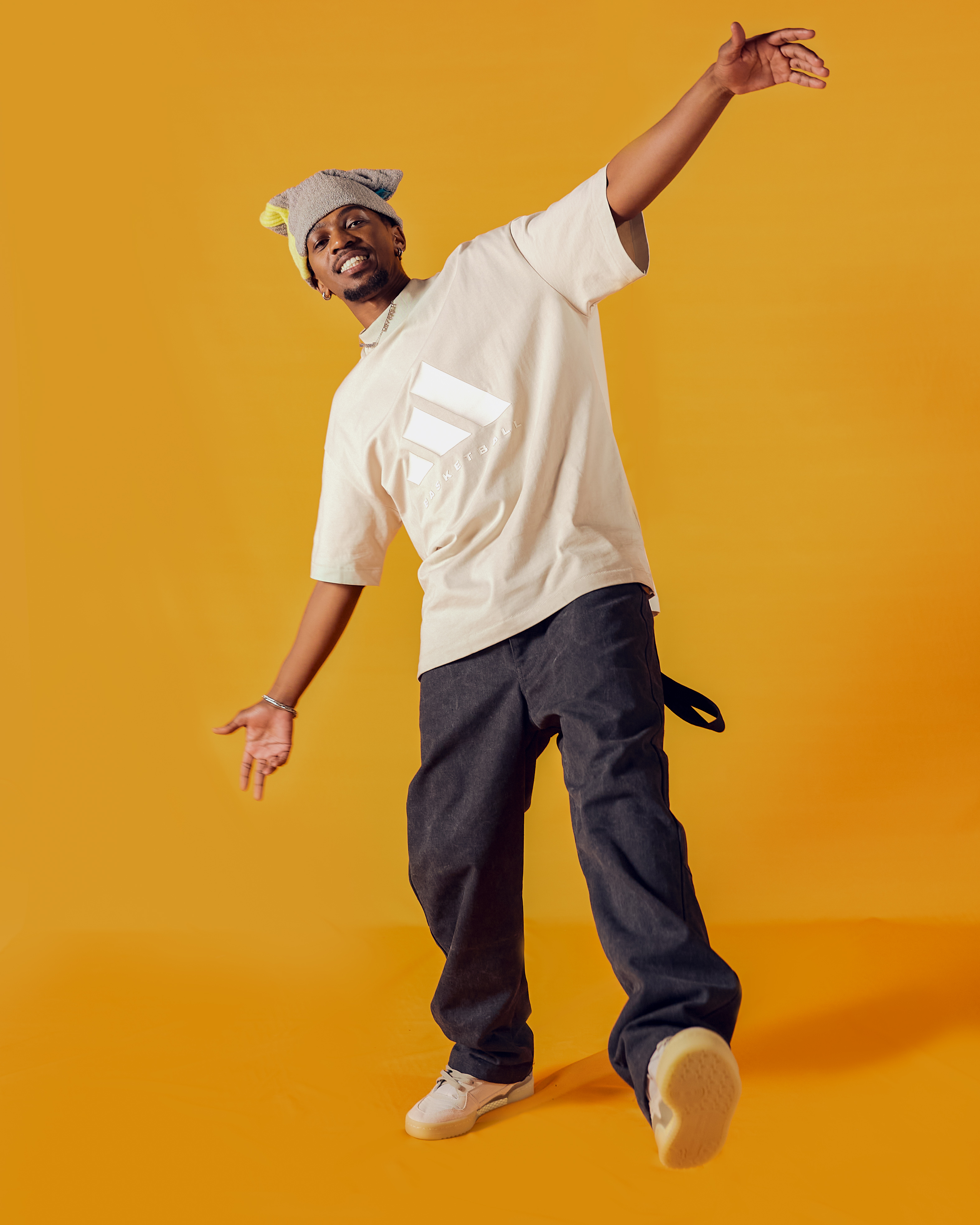 Smiling man in a towel hat, beige shirt, black pants, and white sneakers posing against orange background.