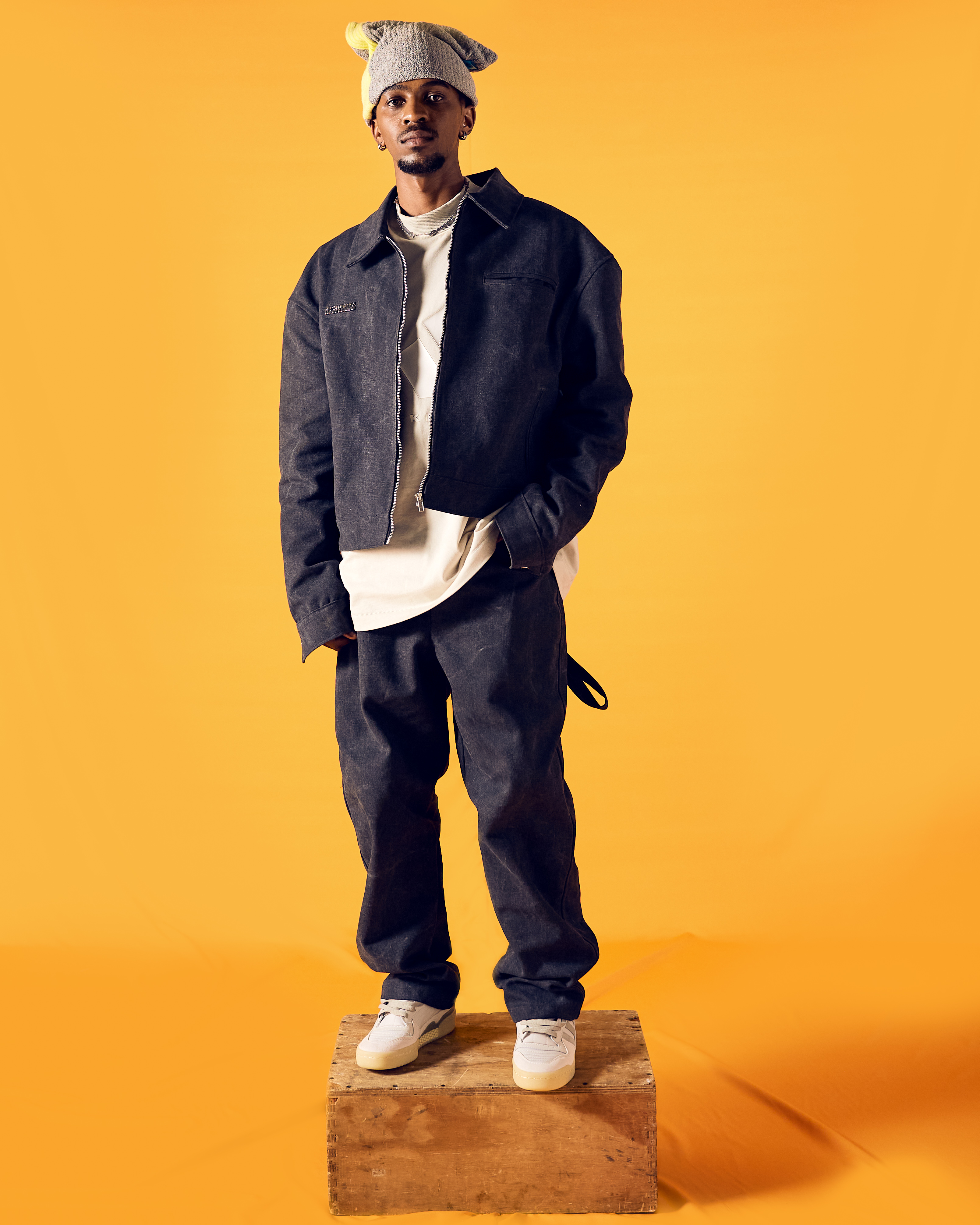 Man wearing a gray jacket, pants, and hat standing on a wooden box against an orange background.
