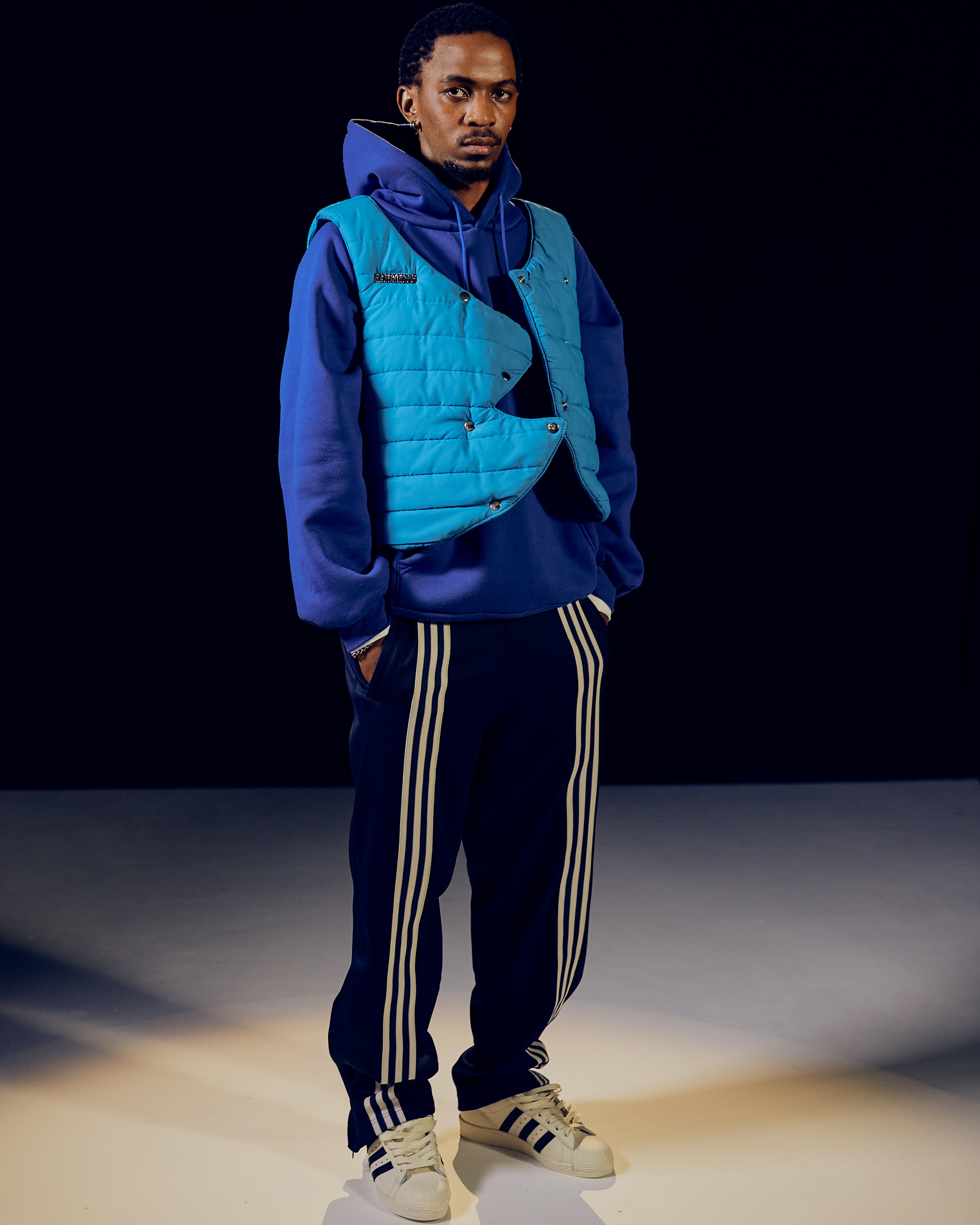 Man wearing blue hoodie, teal vest, striped track pants, and white sneakers standing against dark background.