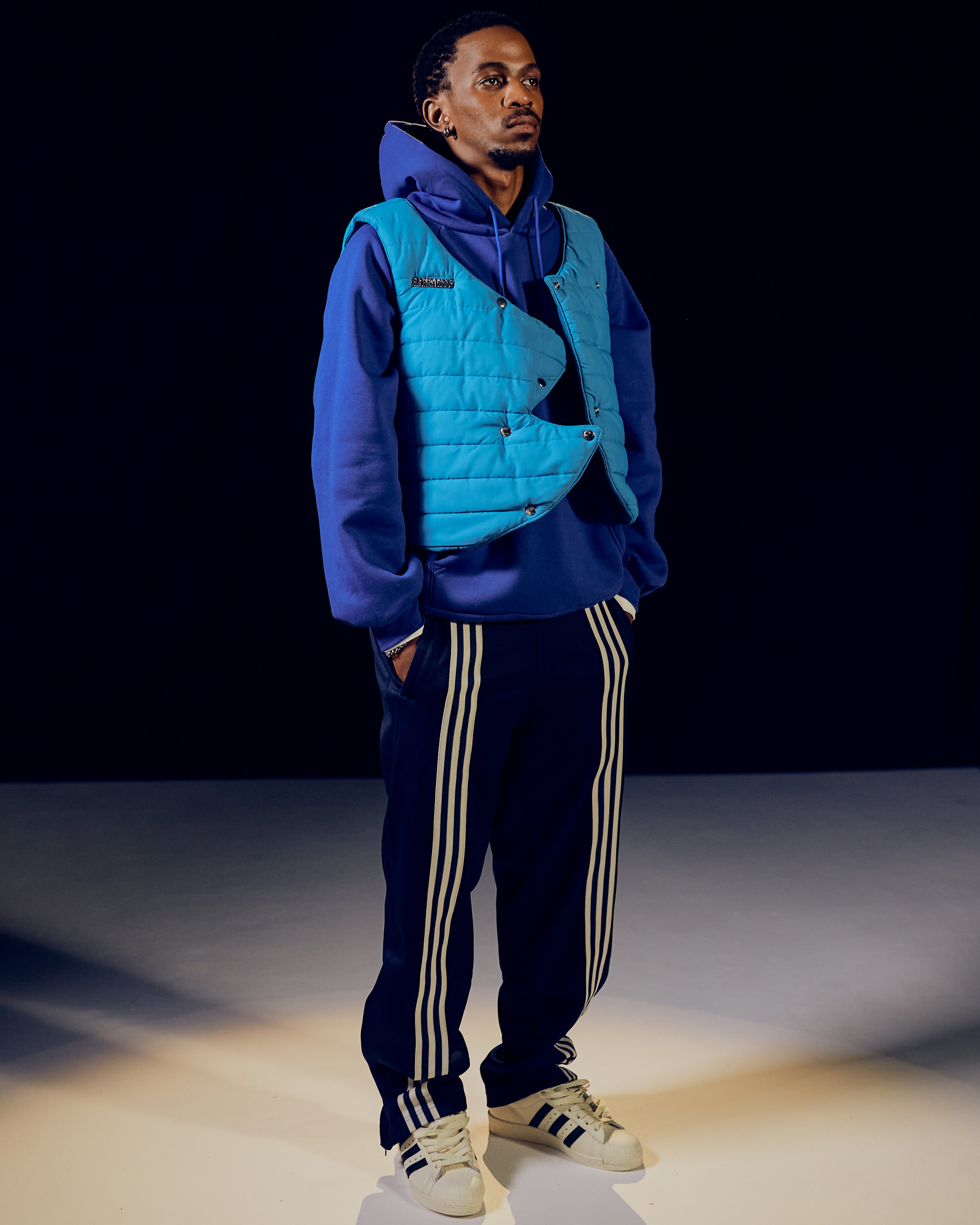 Man in blue hoodie, teal vest, navy track pants with white stripes, and white sneakers standing with hands in pockets.