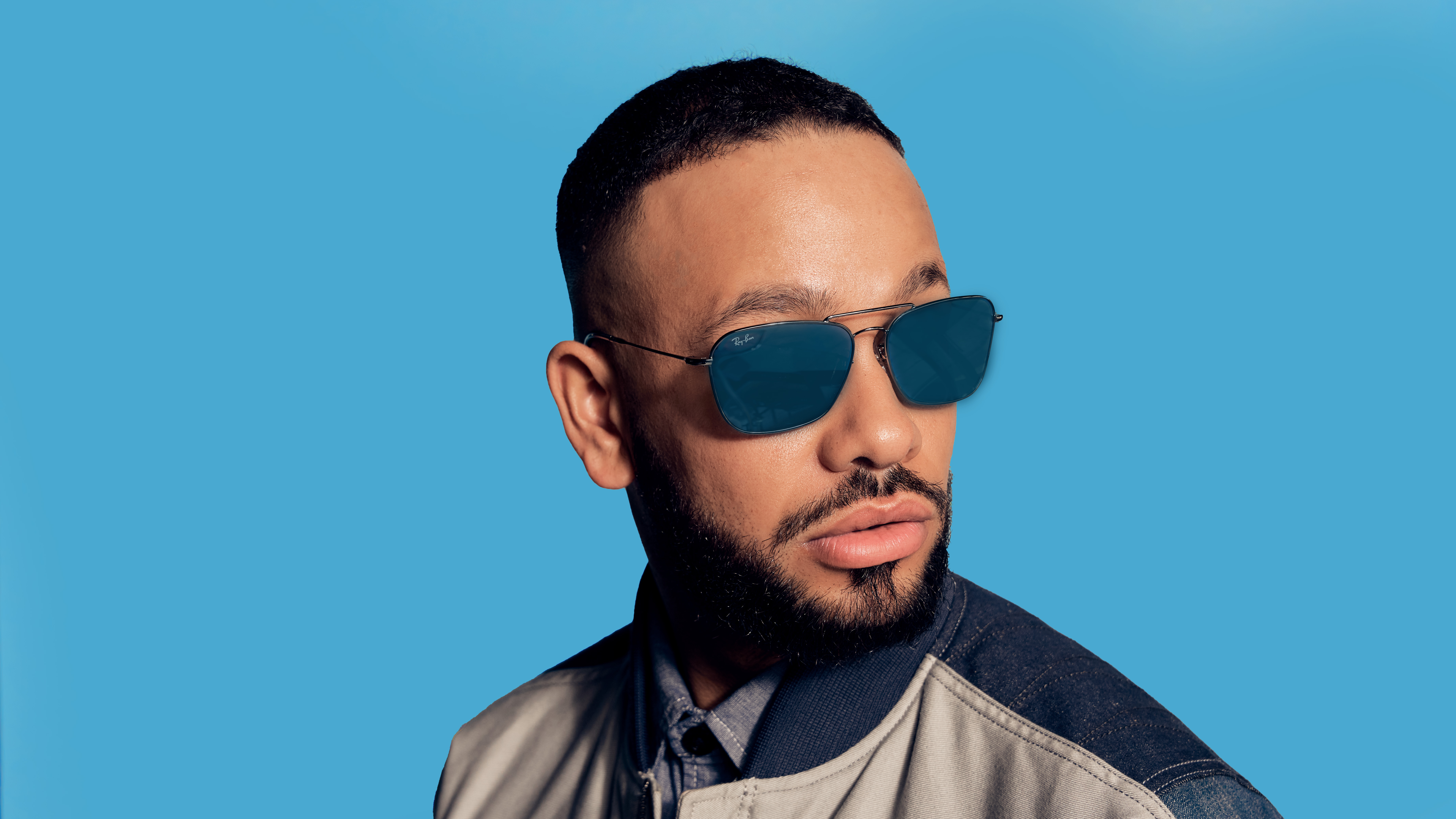 Man with beard wearing dark blue sunglasses and a jacket against a blue background.