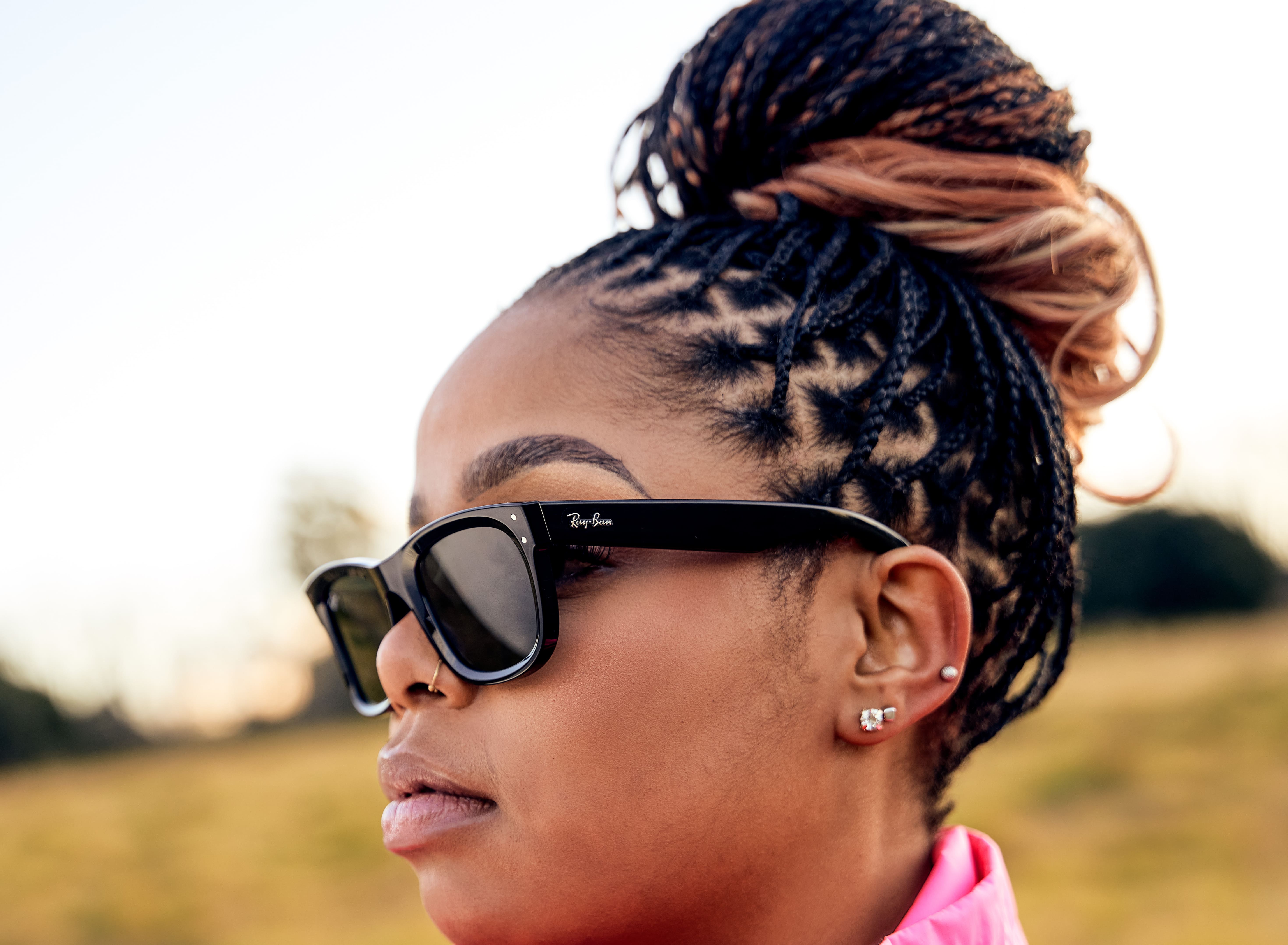 Side profile of a woman wearing black Ray-Ban sunglasses outdoors with braided hair in a bun.