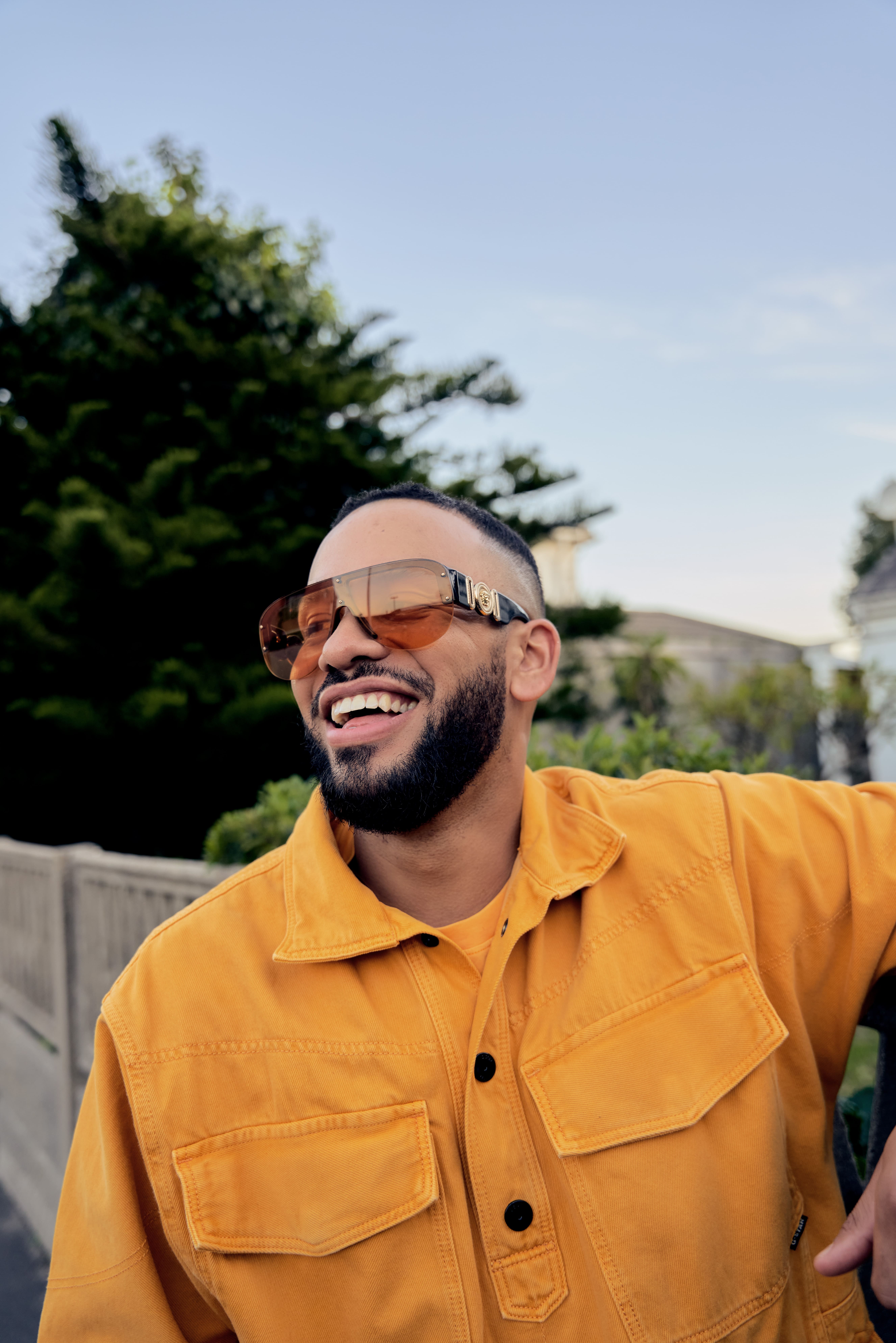 Smiling man with beard wearing orange jacket and large brown sunglasses outdoors near greenery.