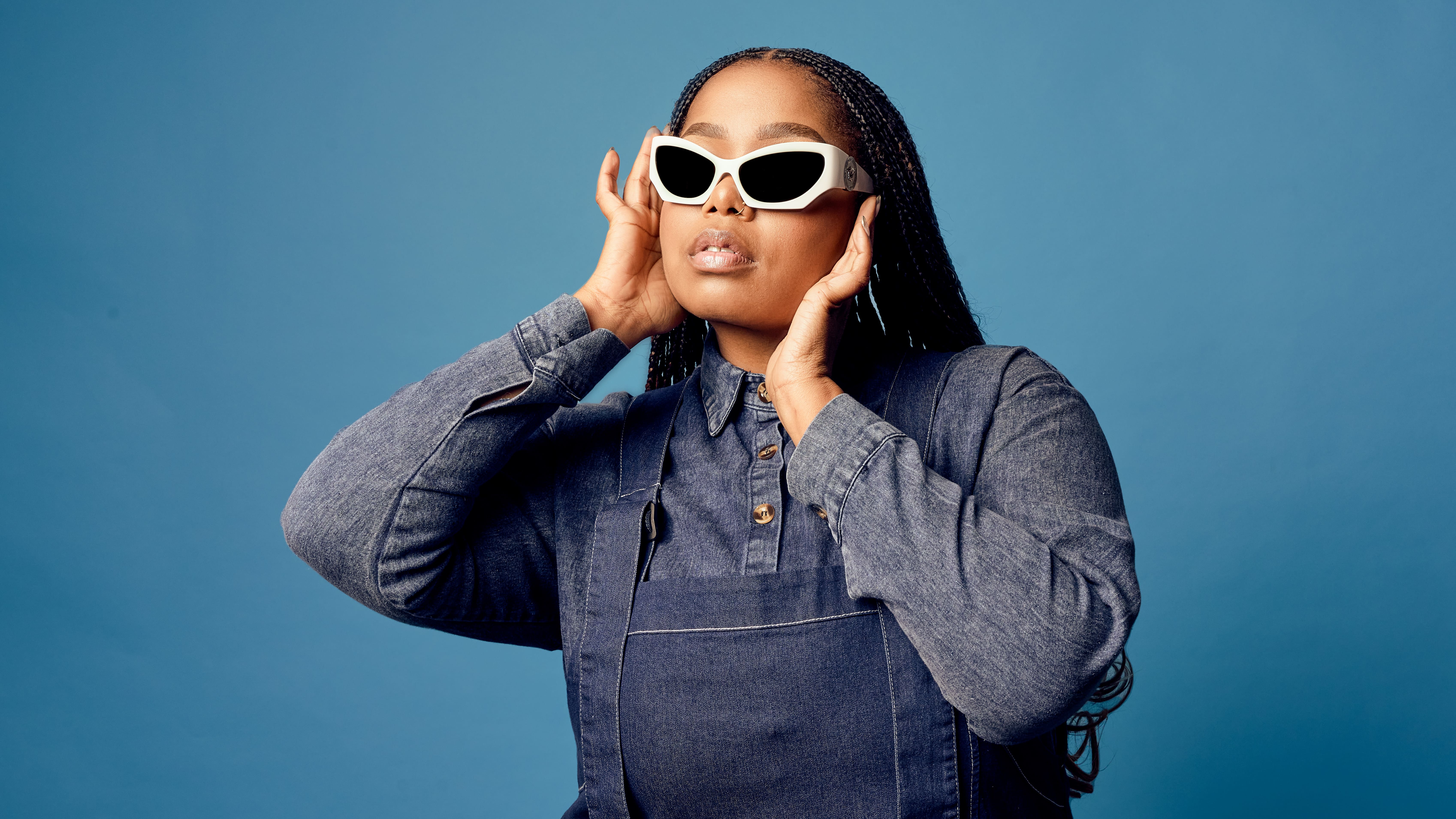 Woman wearing white cat-eye sunglasses and denim outfit posing against a blue background.