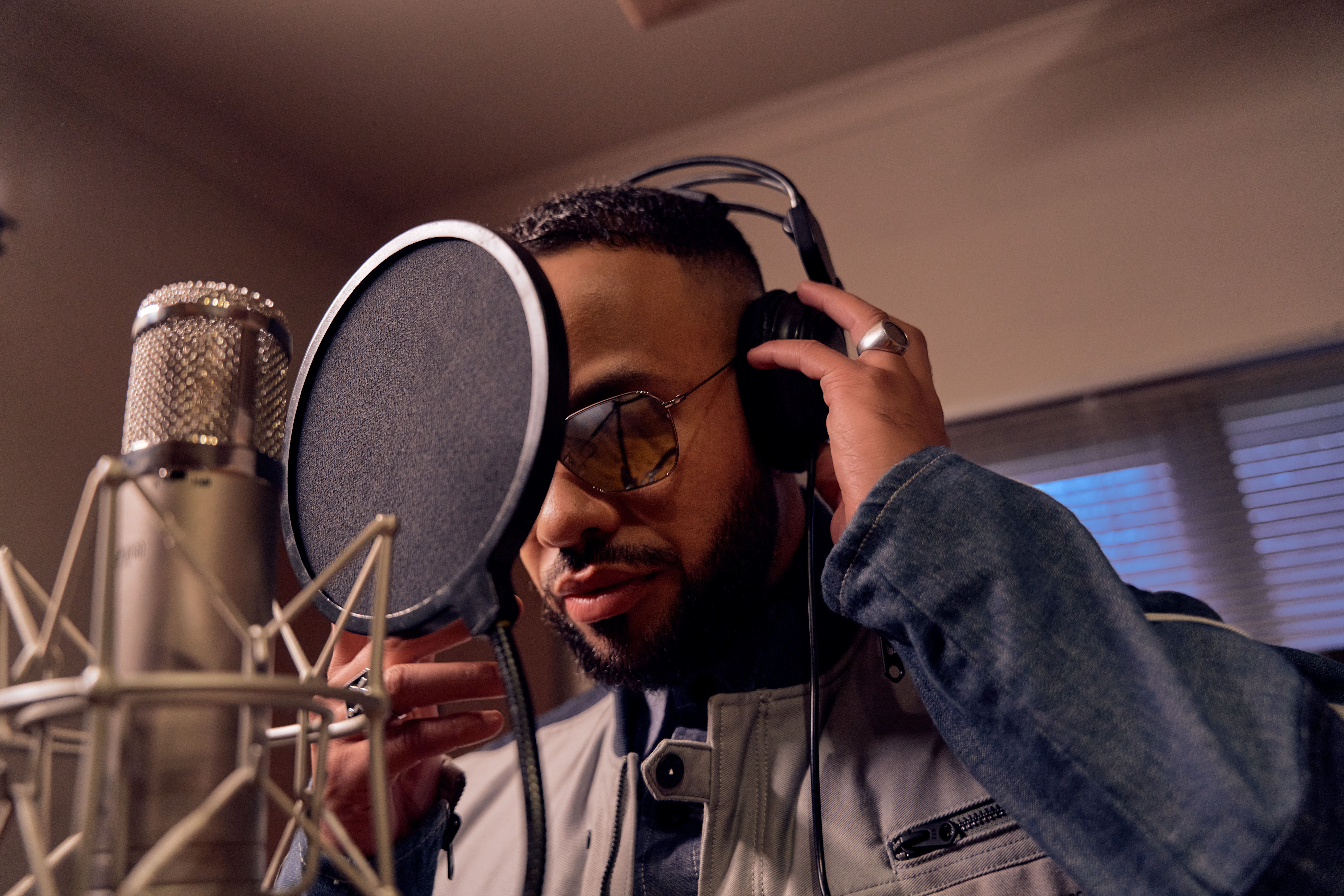 Man wearing sunglasses and headphones recording vocals in front of a studio microphone pop filter.